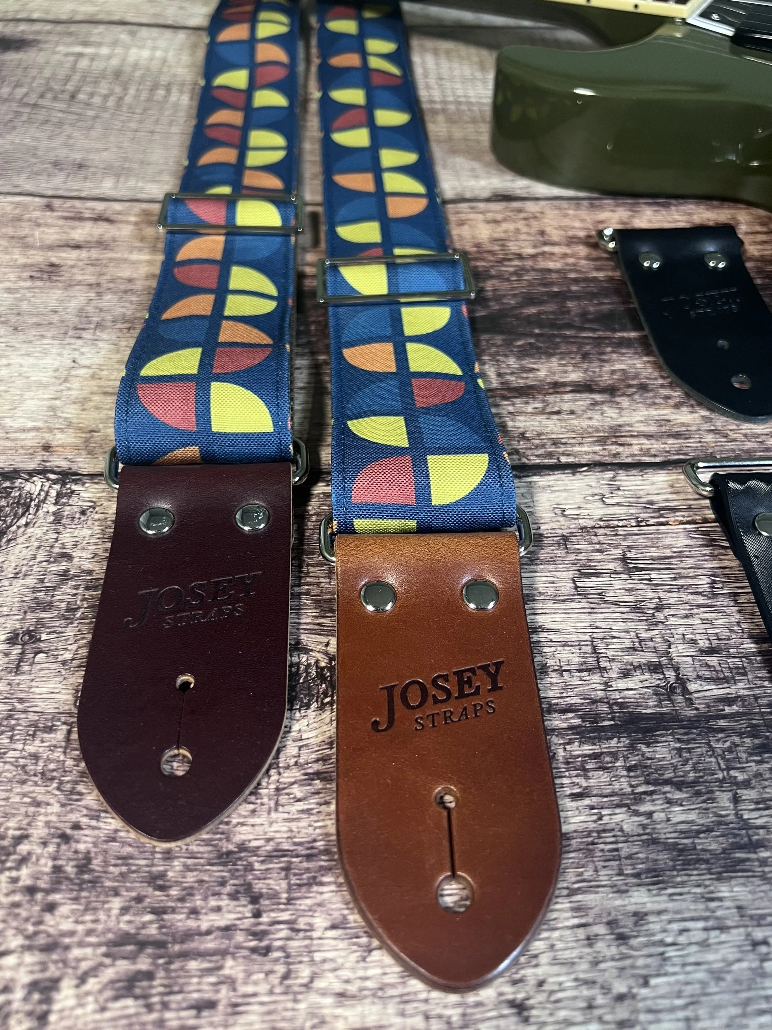 Two guitar straps in navy blue, with geometric circle print in mustard yellow, coral and steel blue laid flat on a wood background