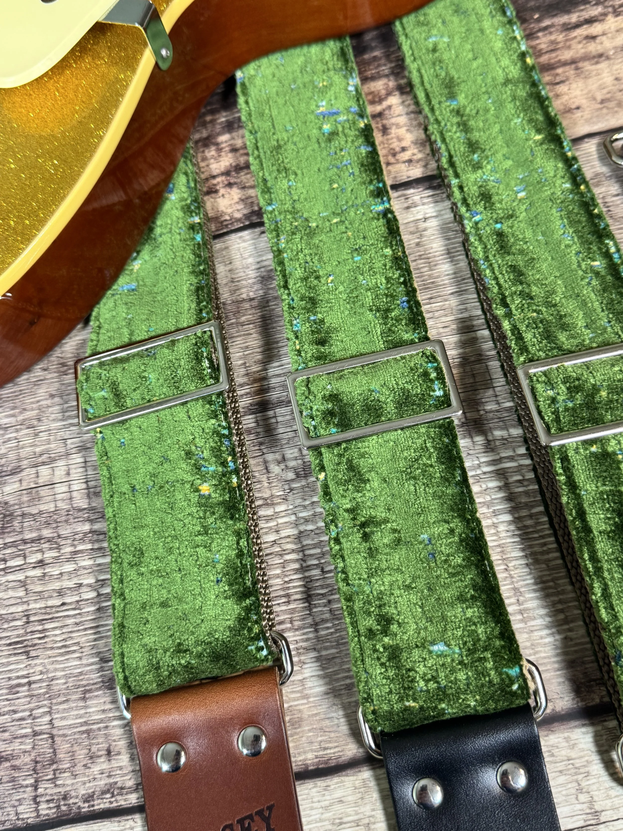Handmade green velvety guitar strap with stamped leather ends laid on weathered wood next to a gold sparkle electric guitar — The Grouch by Josey Straps