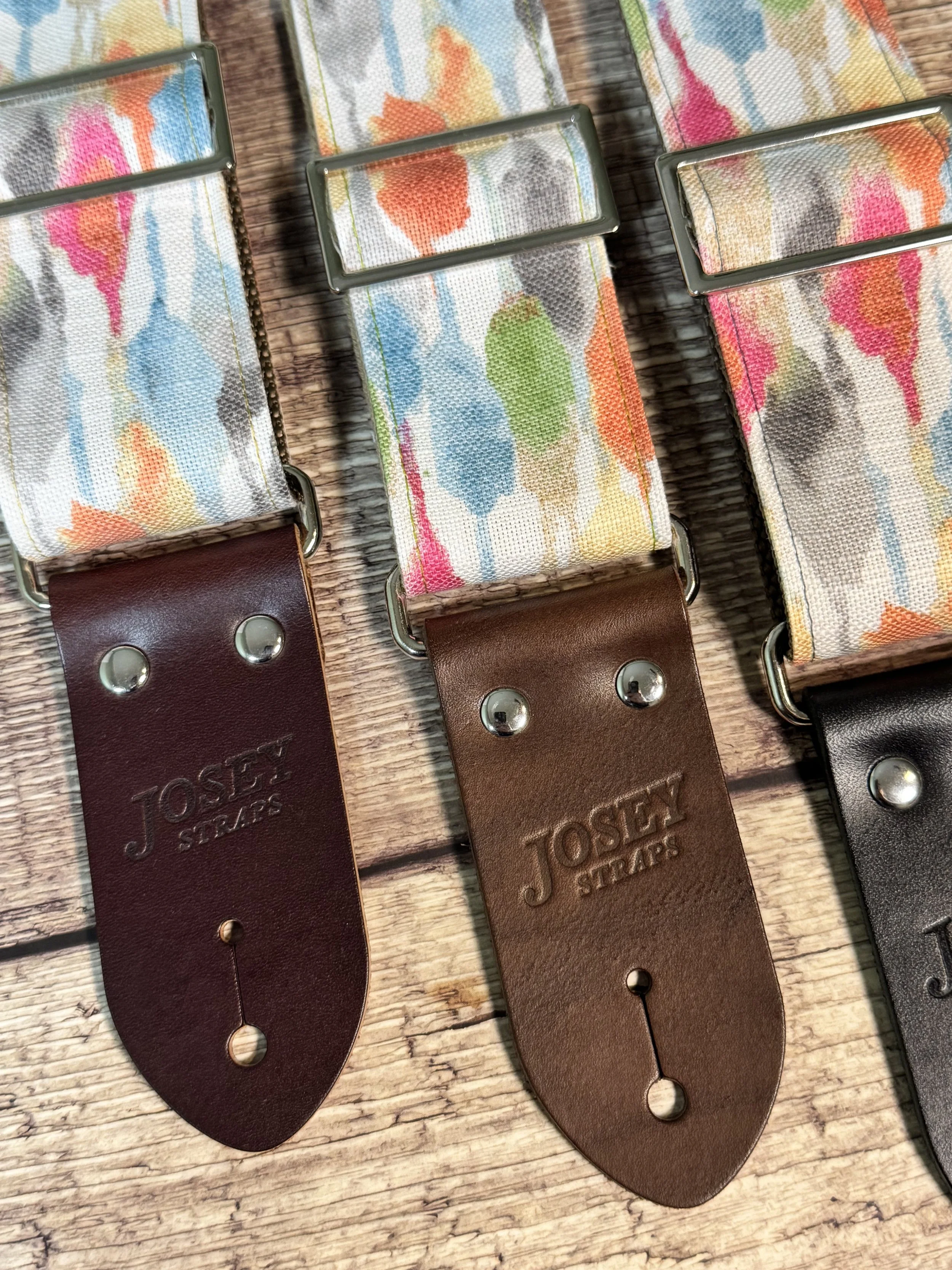 Josey Straps guitar straps in the April colorway — a watercolor print of coral, blue, sage, and amber on cream — displayed on a wood surface.
