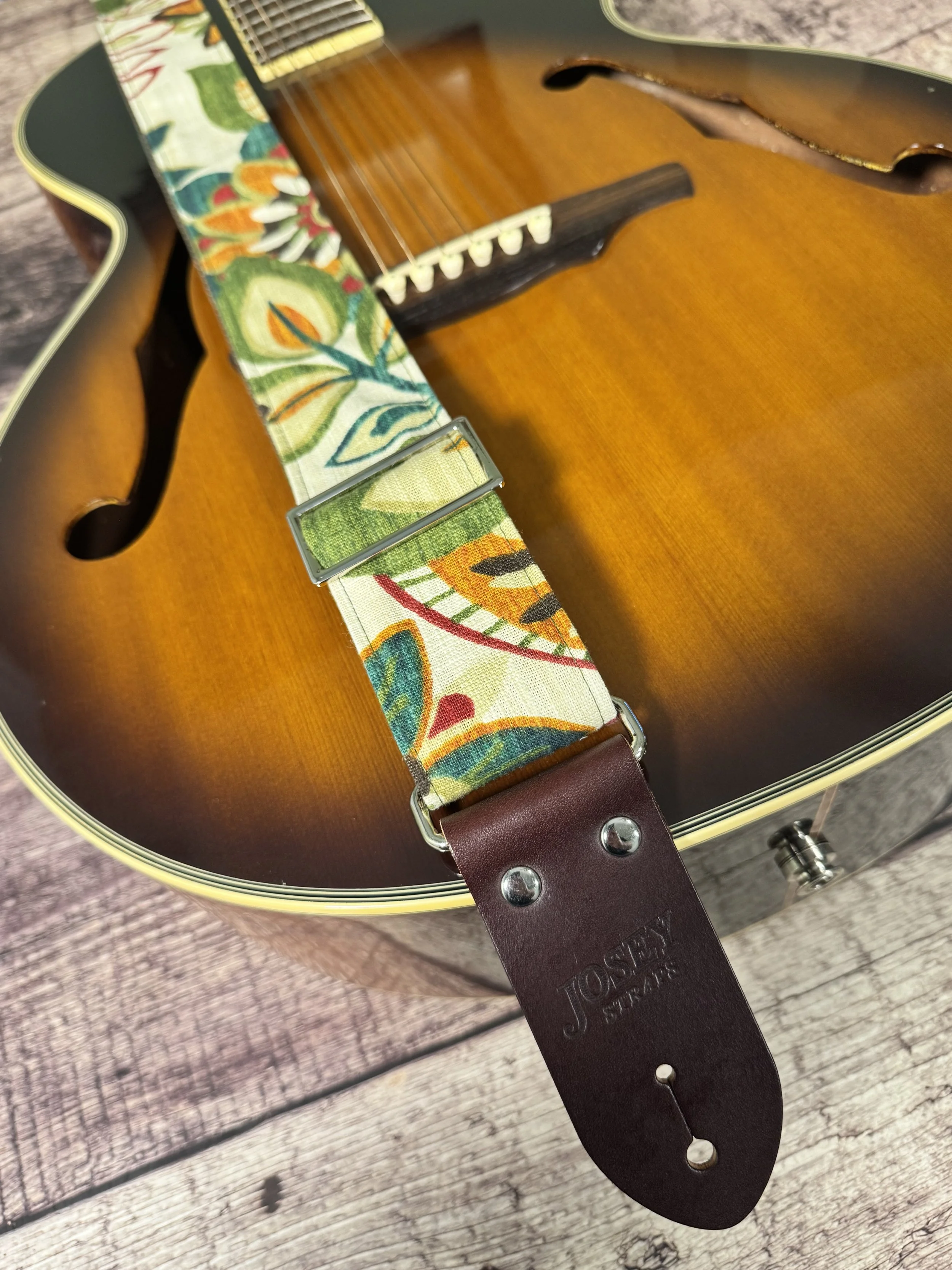 Handmade guitar strap in cream botanical fabric with amber, teal, burgundy, and forest green floral print, laid flat on a wood surface on an acoustic guitar.