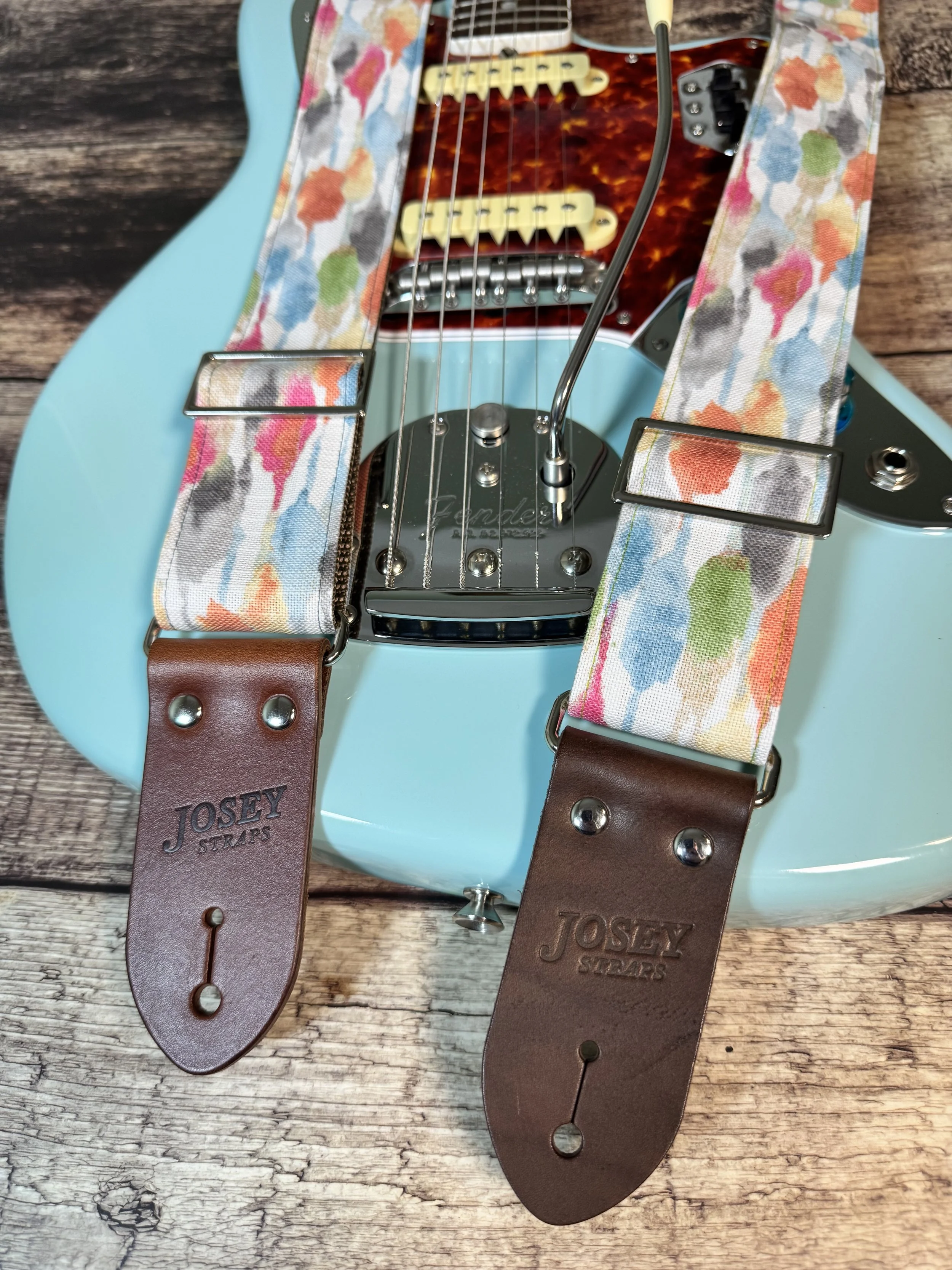 Josey Straps guitar straps in the April colorway — a watercolor print of coral, blue, sage, and amber on cream — displayed on a wood surface with a daphne blue Fender guitar.