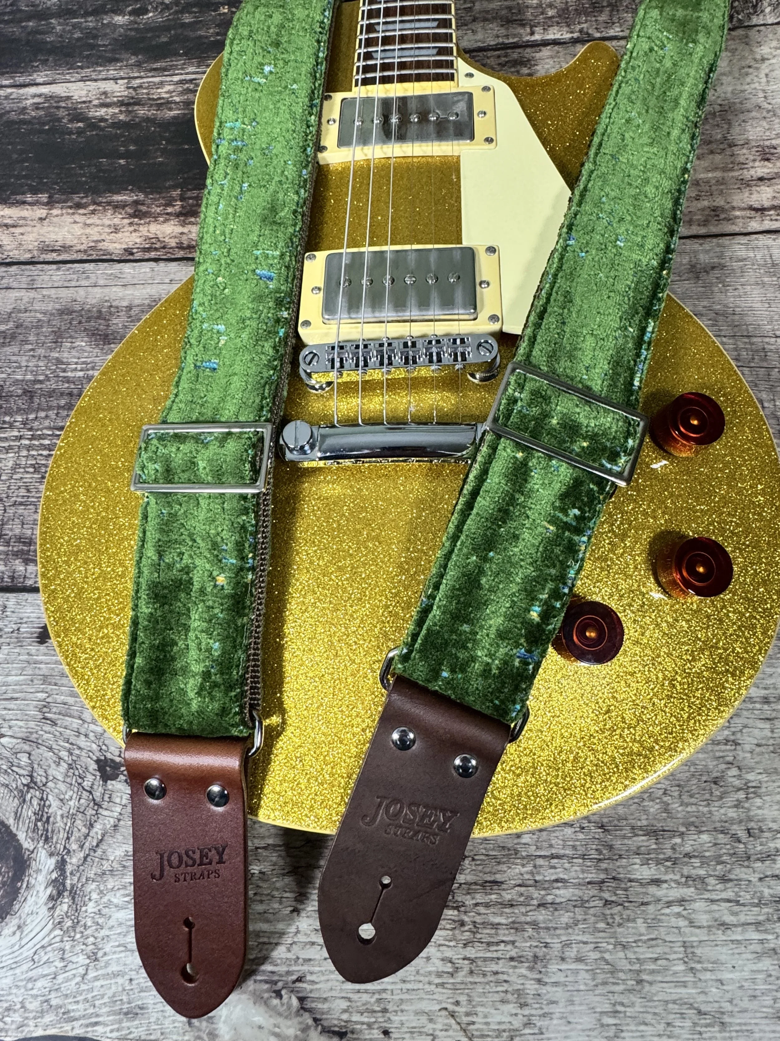 Handmade green velvety guitar strap with stamped leather ends laid on weathered wood next to a gold sparkle electric guitar — The Grouch by Josey Straps
