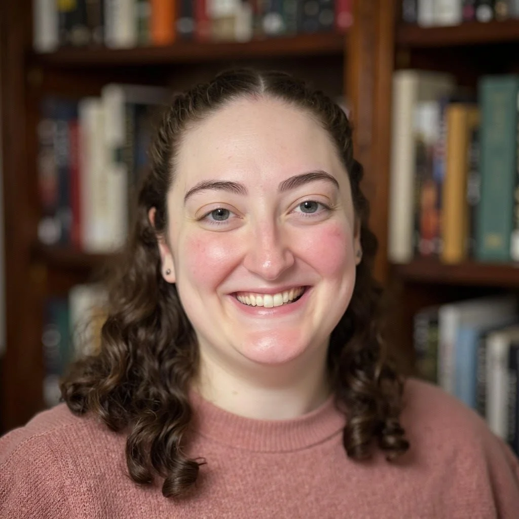 A woman with curly brown hair smiling in front of a bookshelf filled with books.