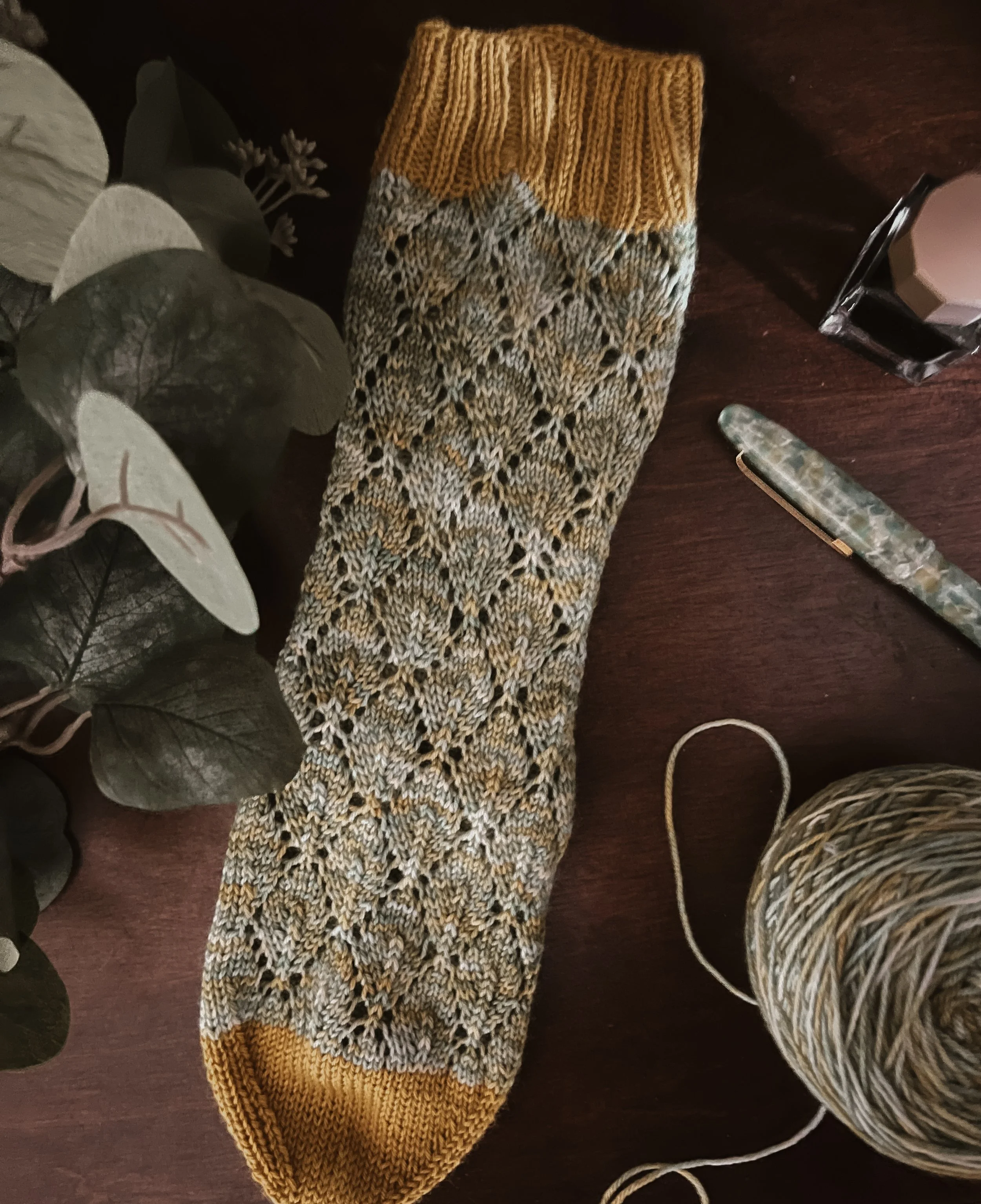A partially finished knitted sock in yellow, gray, and black yarn, placed on a dark wooden surface next to a ball of yarn, a metallic green knitting needle, a white candle in a glass holder, and some green leaves.