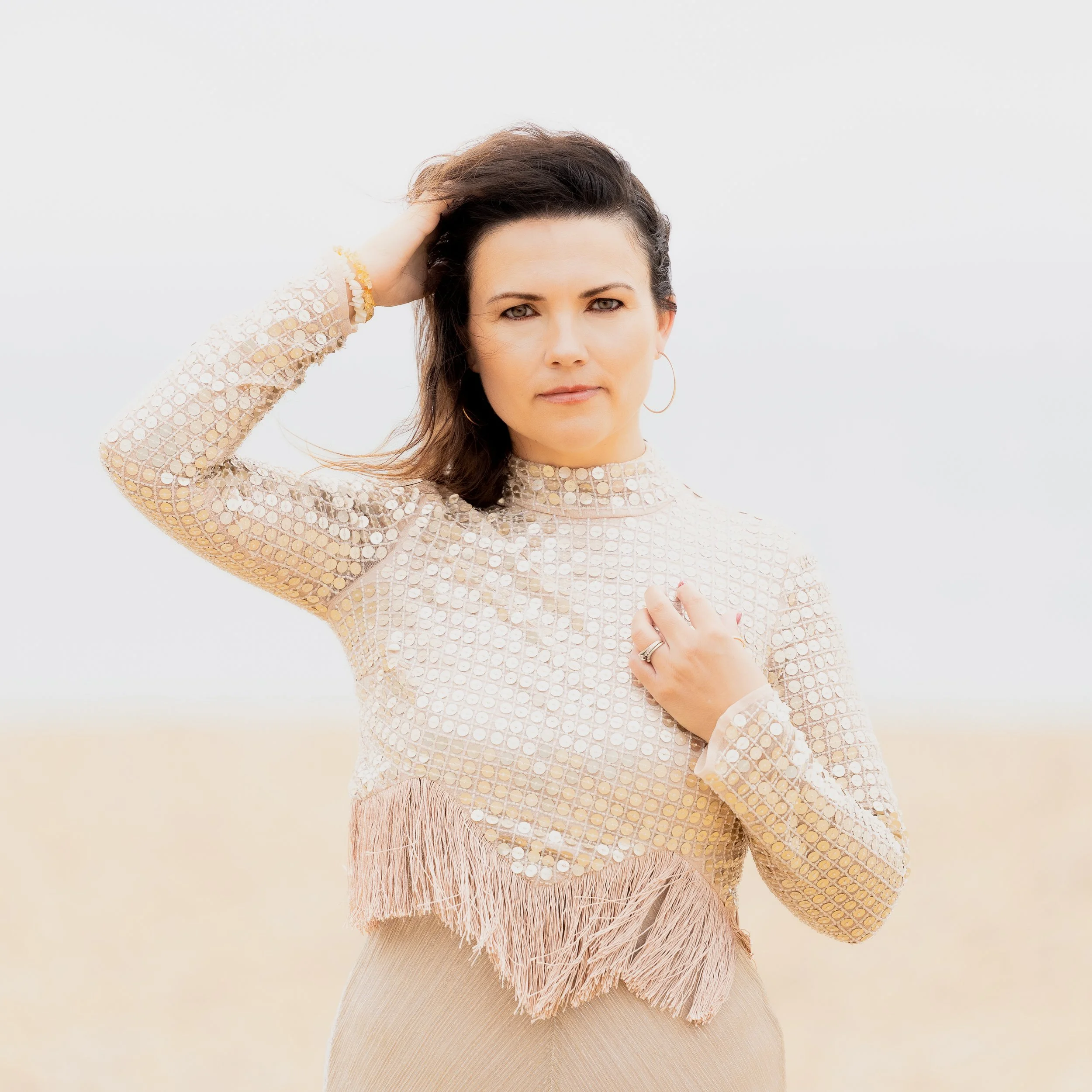 A woman with dark brown hair and light skin, wearing a beige, long-sleeved, sequined top with fringe and beige pants, stands outdoors against a blurred, light-colored background. She has one hand on her head and the other on her chest, gazing directl