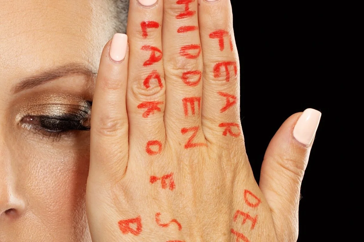 A woman with eye makeup covering one eye, holding her hand up to her face with red words written on her fingers and palm.