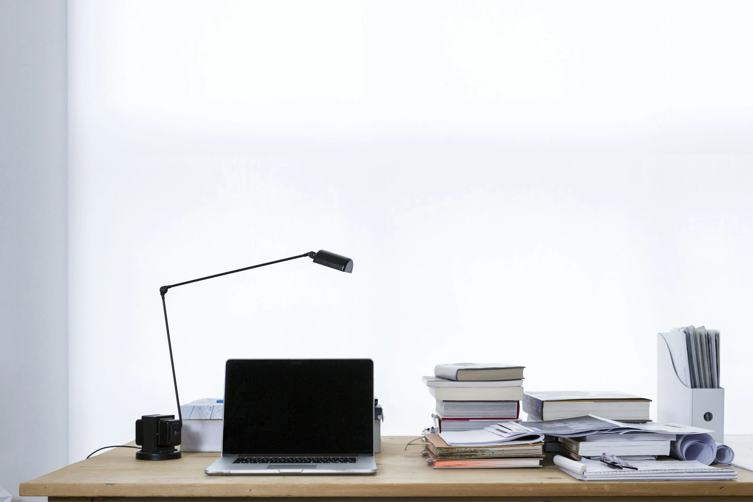 Office desk with a laptop, desk lamp, books, papers, and folders against a white wall.