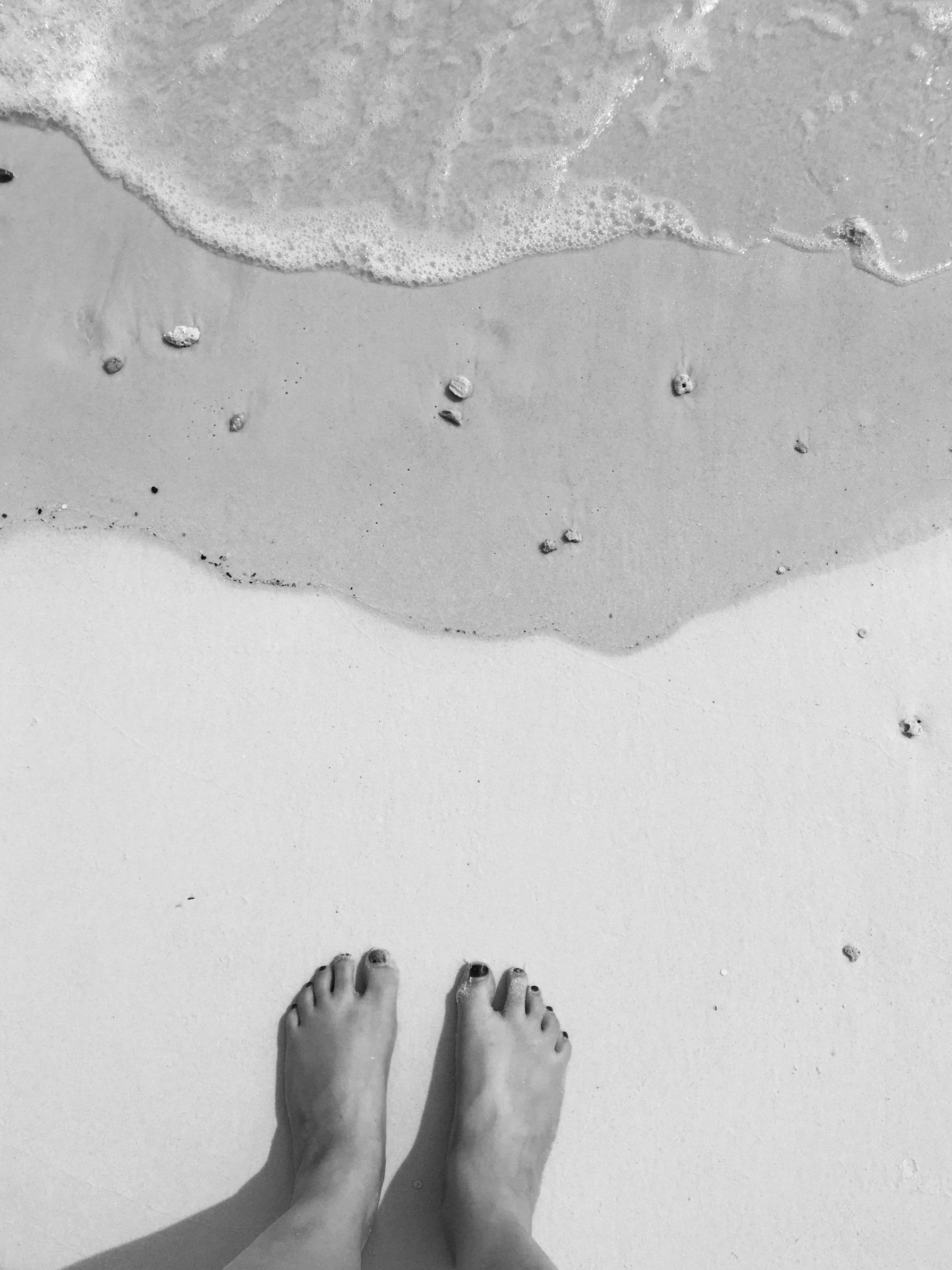 View of a person's feet on a sandy beach with gentle waves at the shoreline.