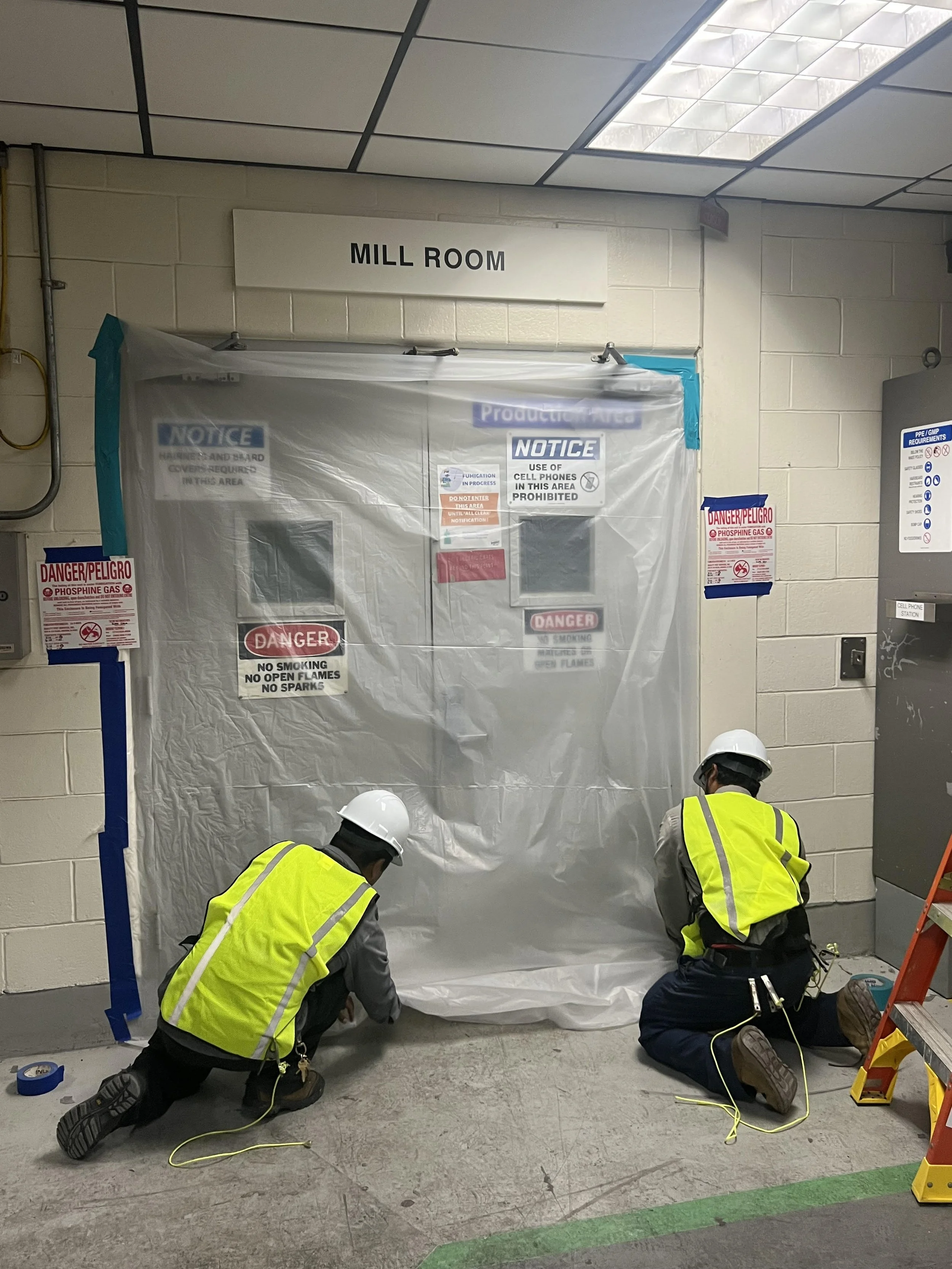 Two workers wearing yellow safety vests and white hard hats working on a plastic-covered area labeled 'Mill Room' with safety signs around them.