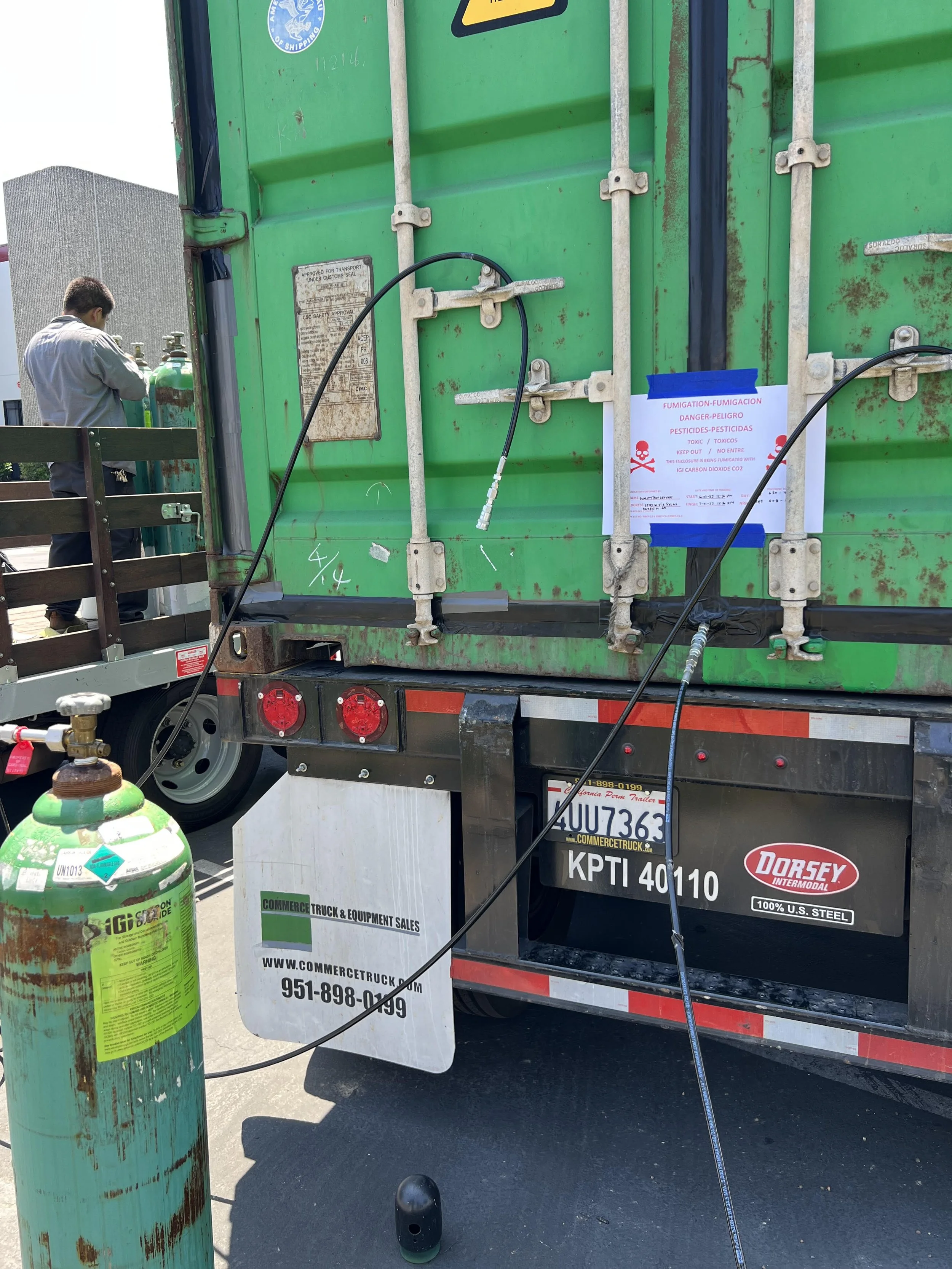 Green shipping container on a truck with a warning sign about fumigation and pesticides, propane tank in the foreground, and a man working nearby.