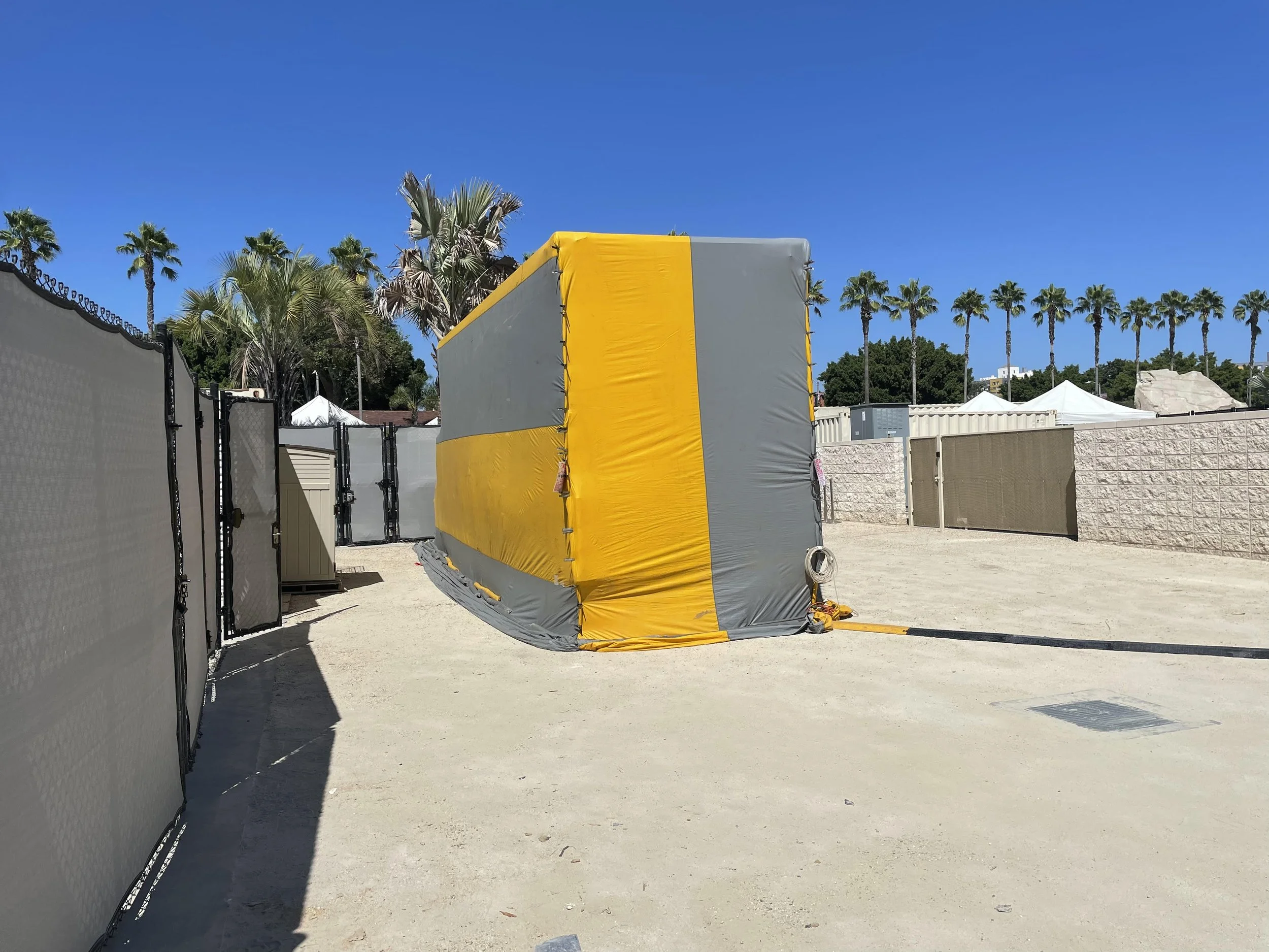 A construction or industrial safety mat structure with gray and yellow padding, situated outdoors with palm trees and a clear blue sky in the background.