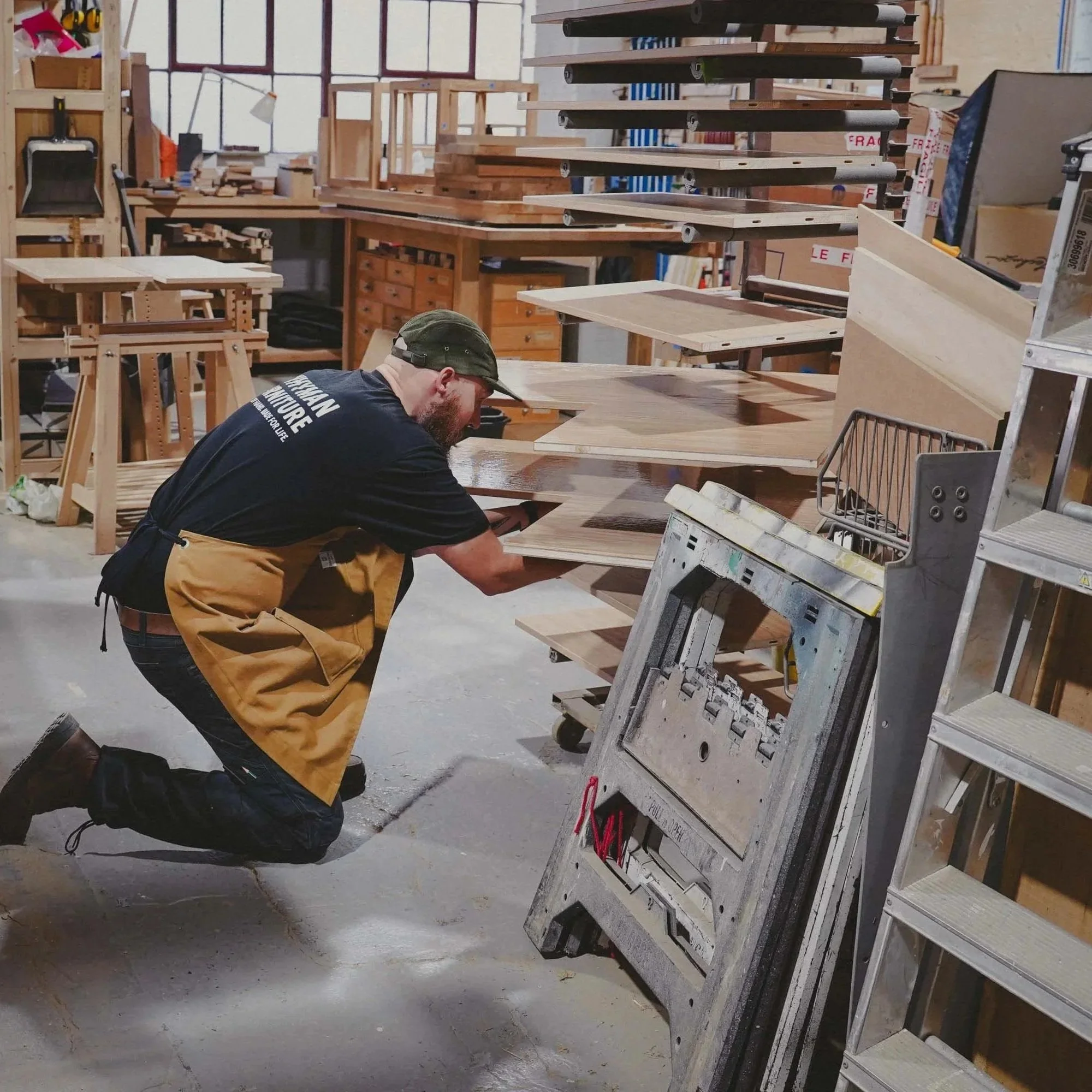 A man in an apron kneels in a woodworking shop, working on wooden boards with tools and equipment around him.