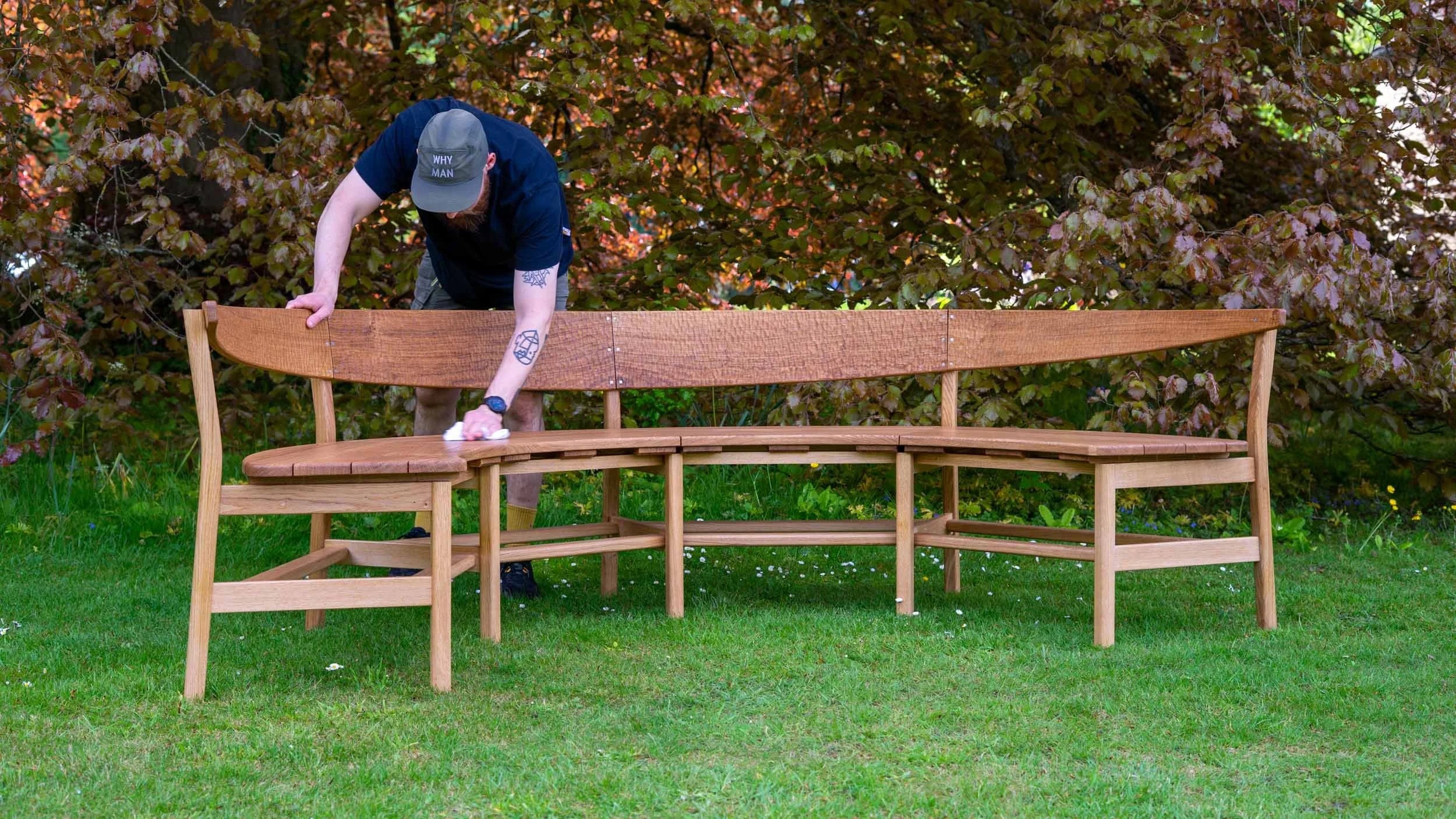 A person in a cap cleans a curved wooden bench on green grass, with leafy trees in the background.