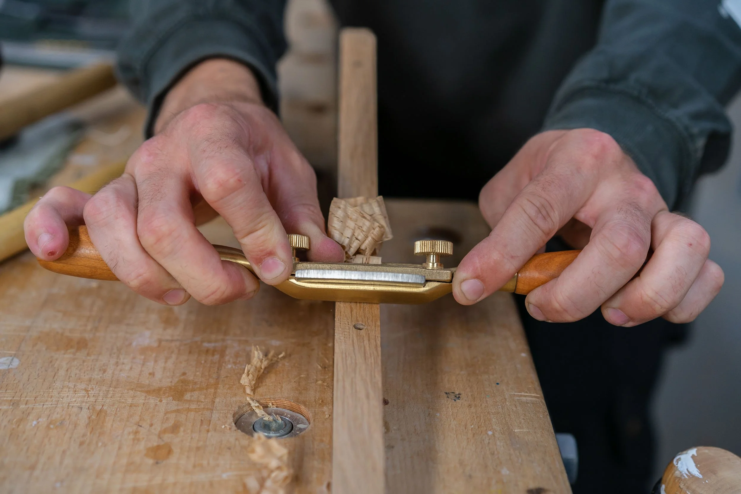 Close-up of hands using a woodworking tool to shave a strip of wood on a workbench, wood shavings visible.