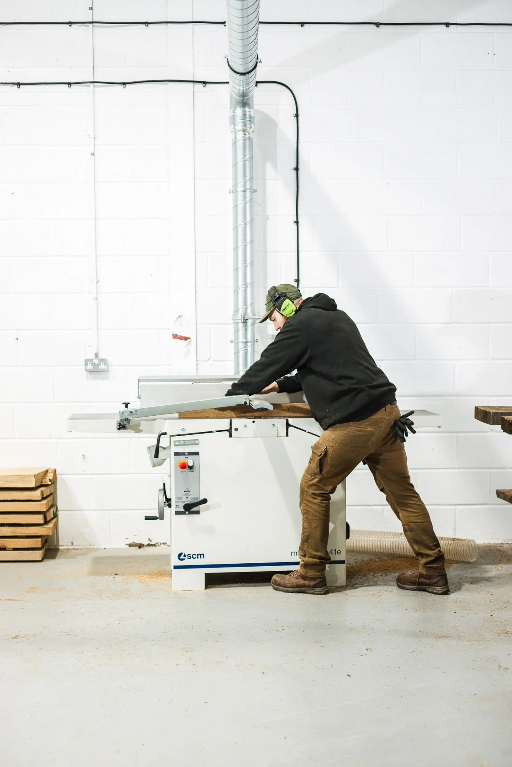 A person wearing ear protection operates a woodworking machine in a bright, clean workshop.