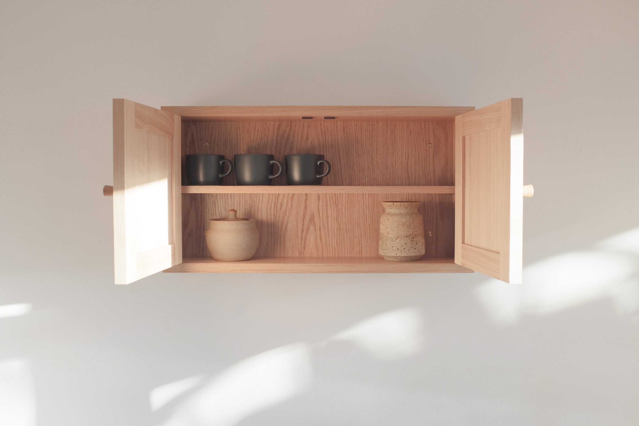Small oak wall mounted cabinet with open doors displaying pottery on shelves inside.