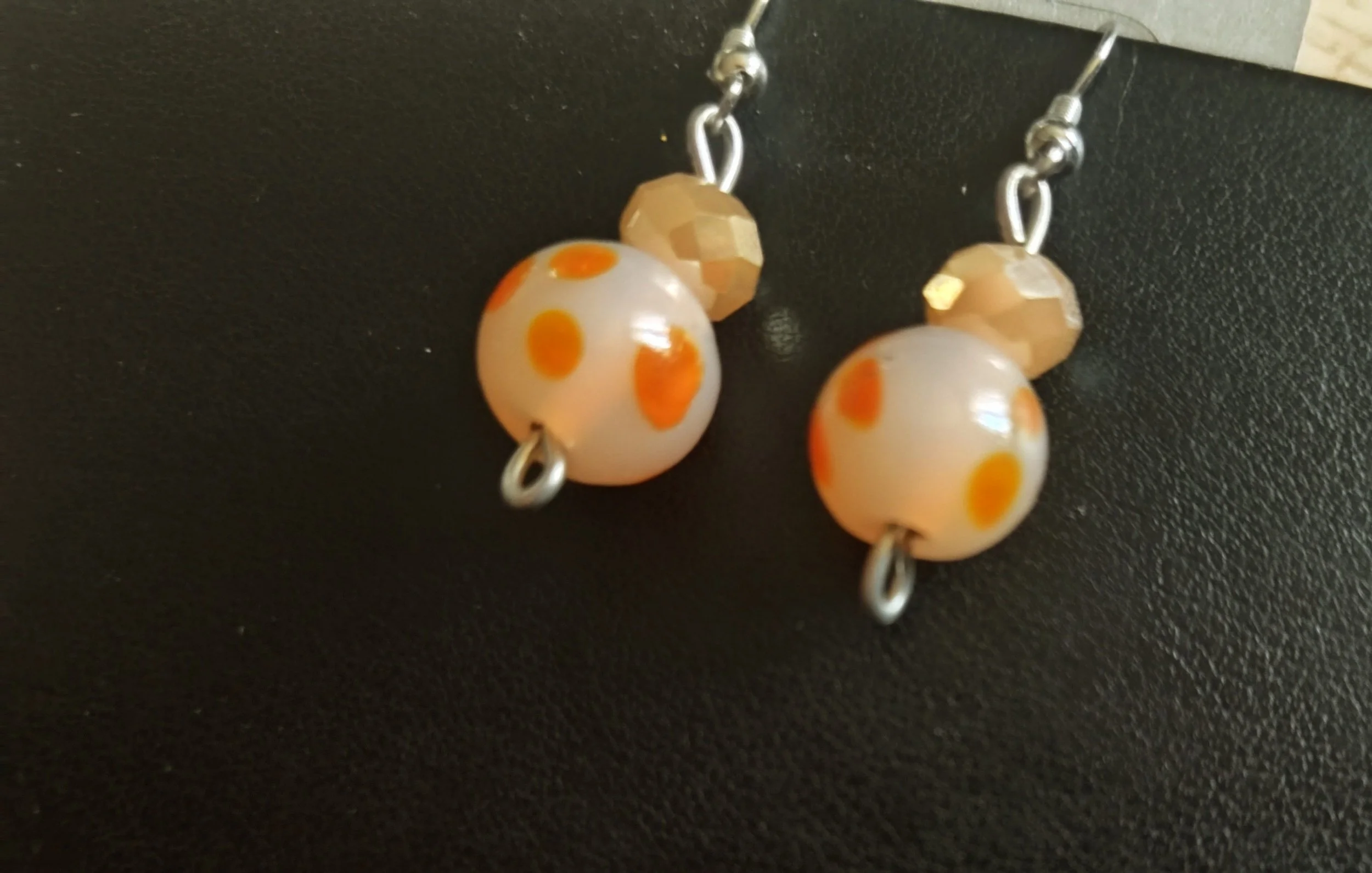 A pair of earrings featuring large white beads with orange polka dots, smaller faceted golden beads, and silver hooks are displayed on a black textured surface.
