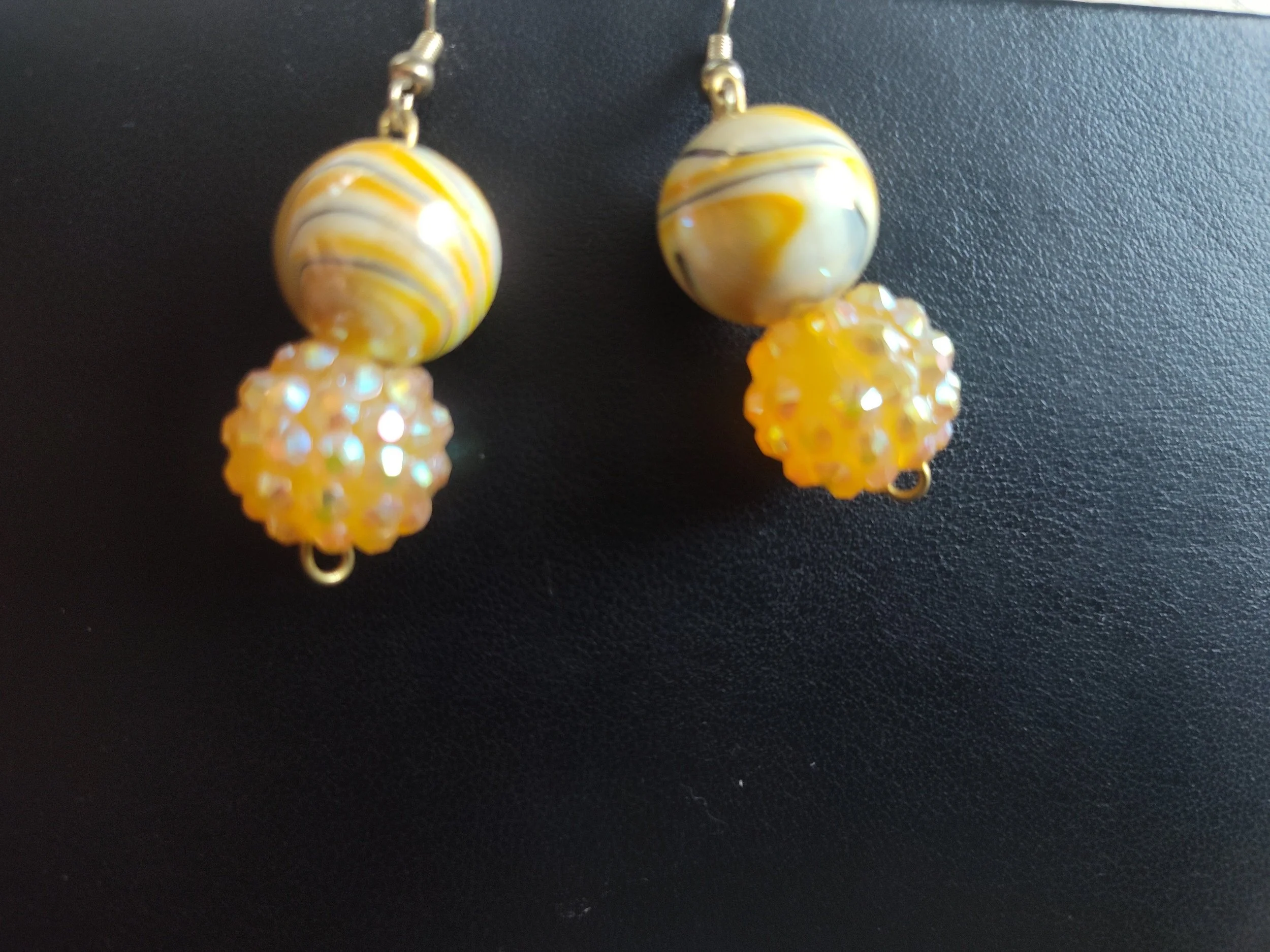 A pair of earrings featuring a large marbled yellow and white bead and a smaller, textured yellow bead with iridescent facets, hanging against a black background.