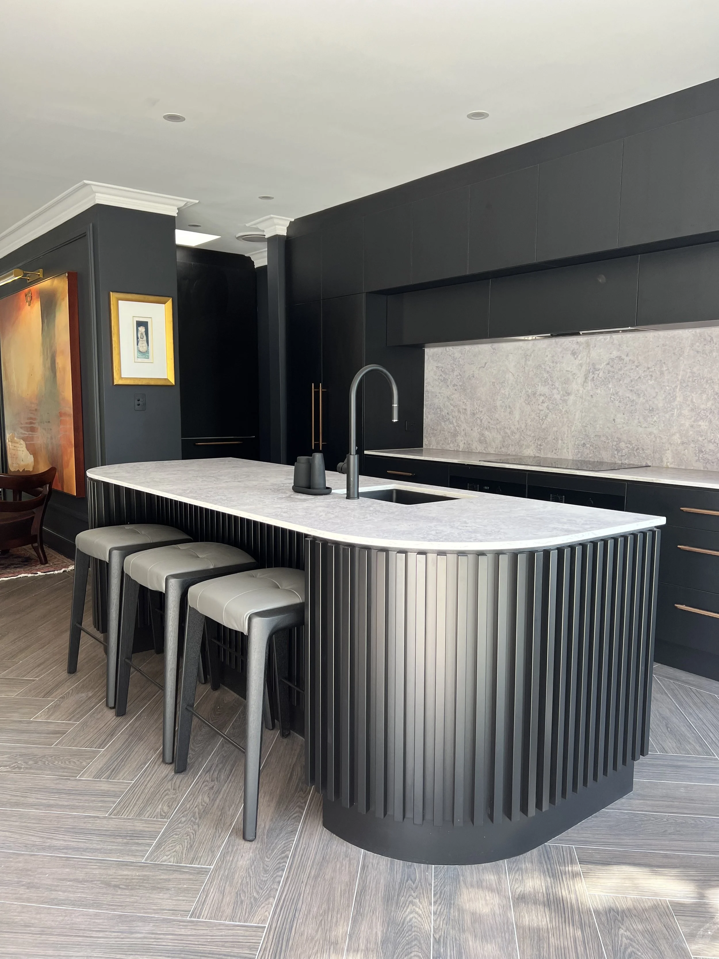 Modern kitchen with black cabinetry, curved island with white marble countertop, black bar stools, and a gray backsplash.