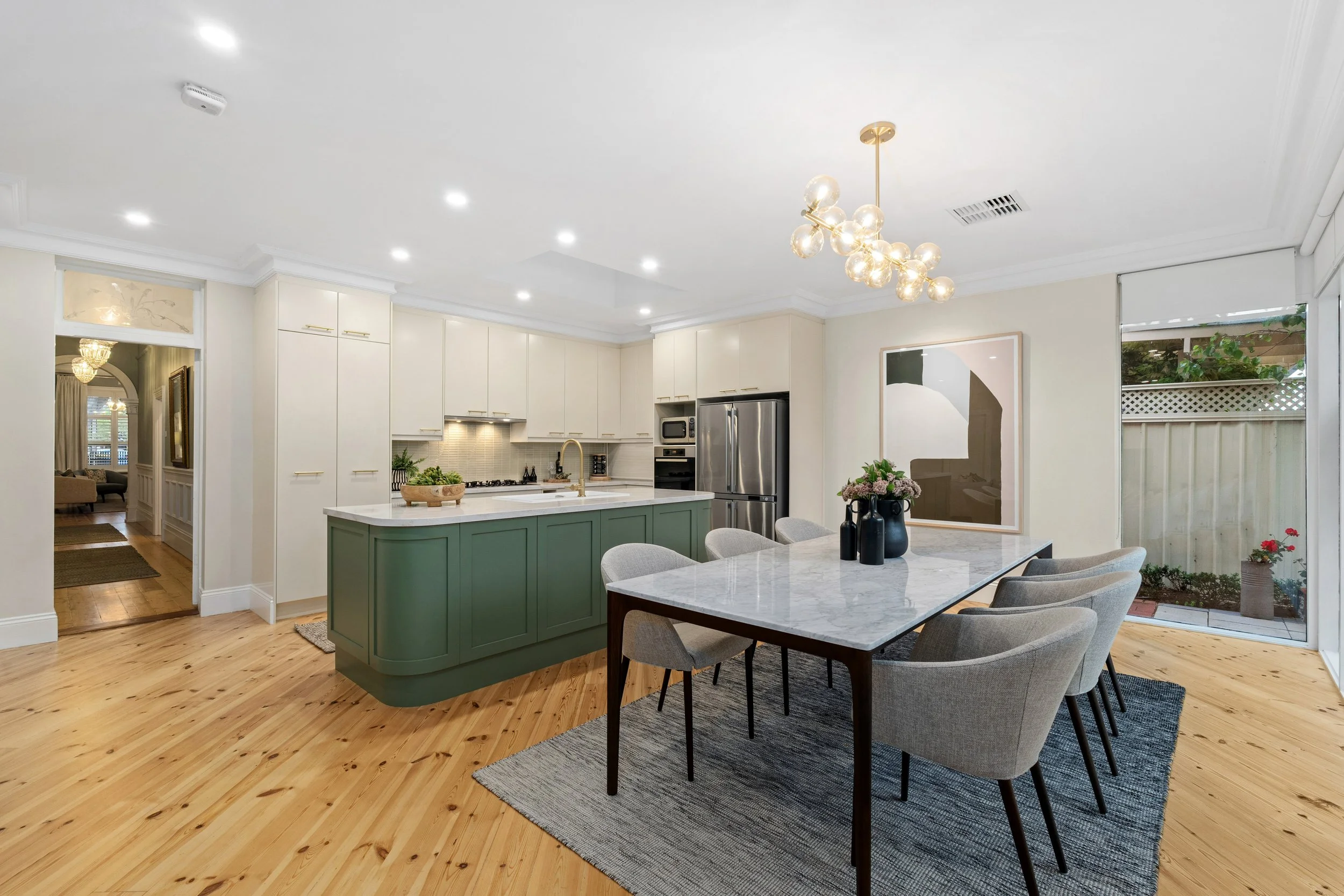 Modern kitchen and dining area with white cabinets, green kitchen island, marble dining table, gray chairs, wooden floor, pendant light, and sliding glass door to the backyard.