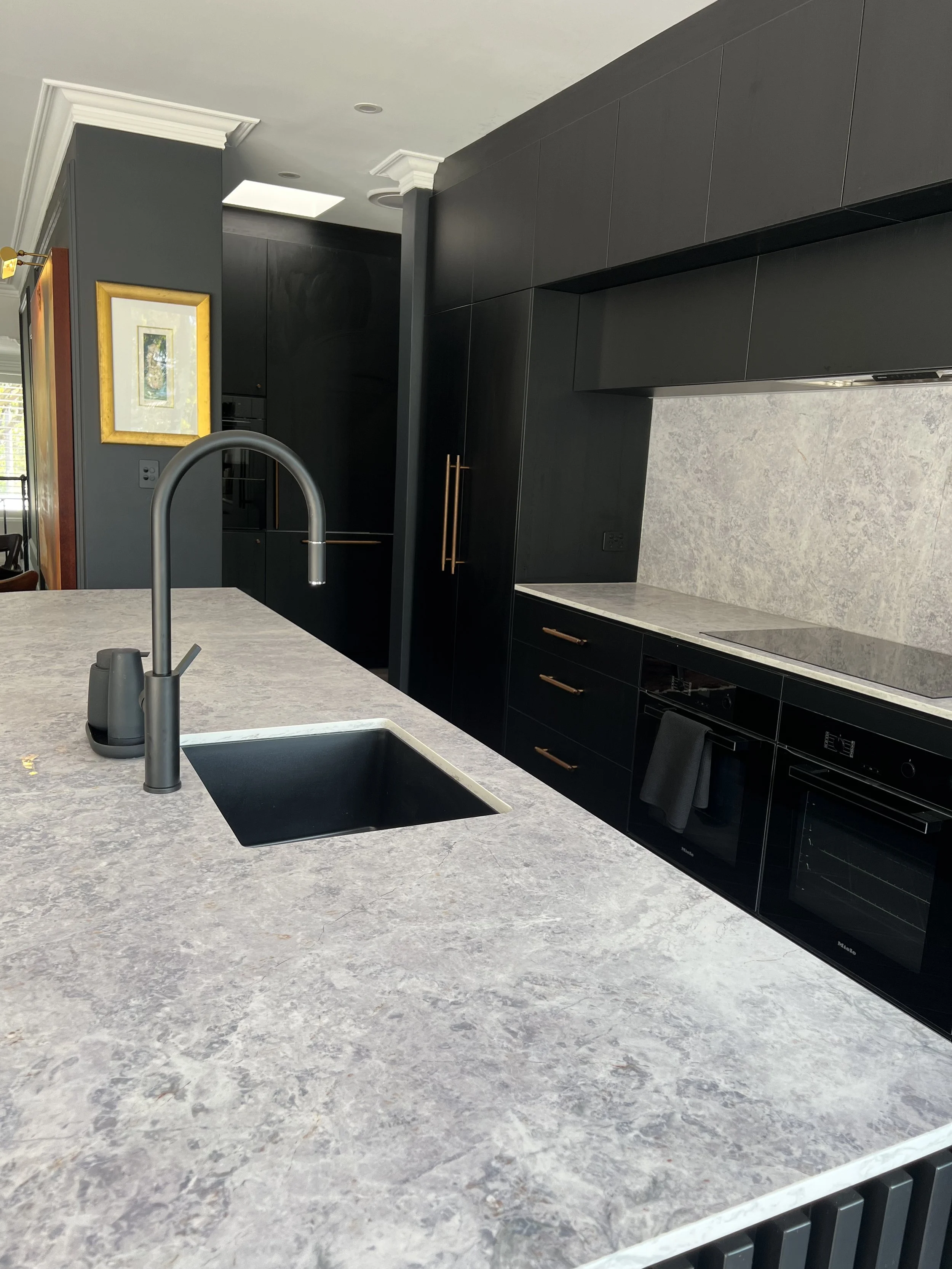 Modern kitchen with dark cabinets, a marble countertop, a black sink, and a gold-framed painting on the wall.