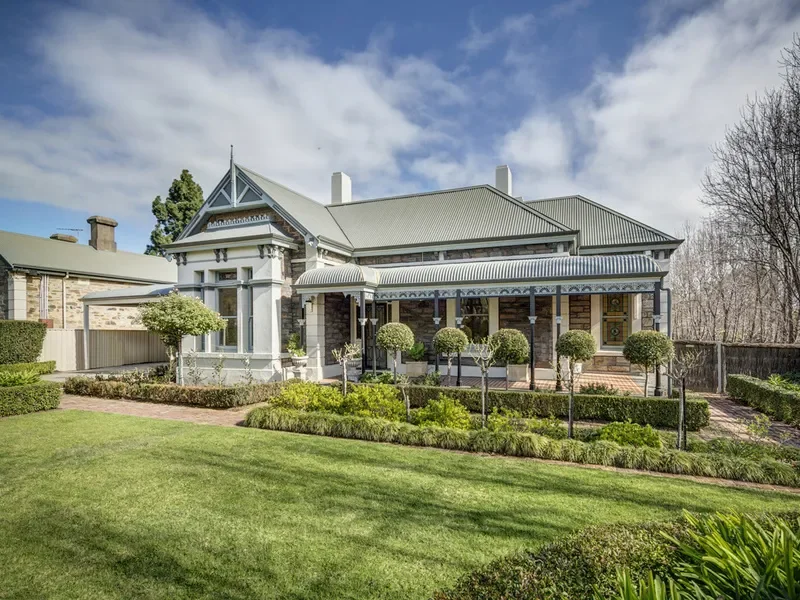 A large historic house with a metal roof, decorative trim, and a front porch with seven evenly spaced trimmed bushes in front. The house is surrounded by a well-maintained lawn and garden, and trees in the background under a partly cloudy sky.