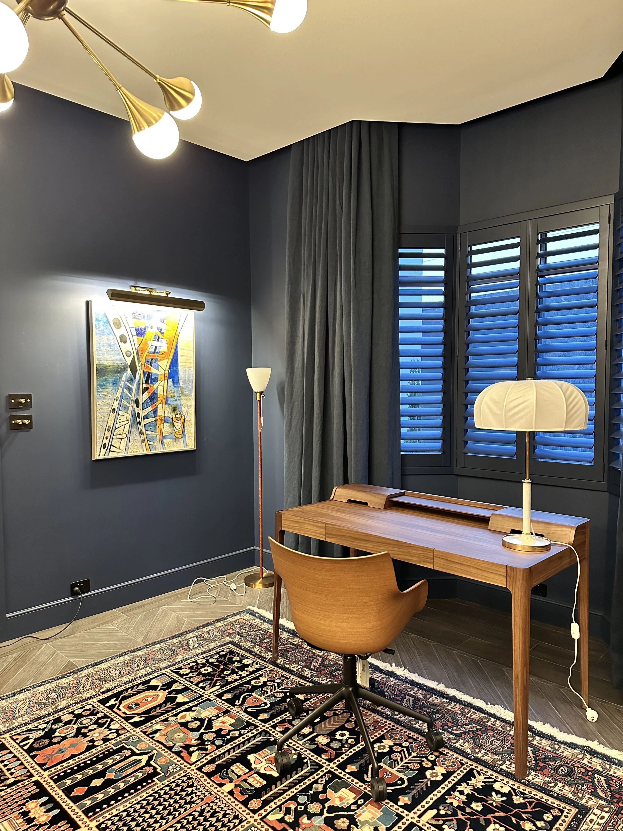 A modern home office with a dark blue wall, a wooden desk, a rolling chair, and a patterned rug. There's a large window with shutters and curtains, a desk lamp, a floor lamp, and a ceiling light fixture.