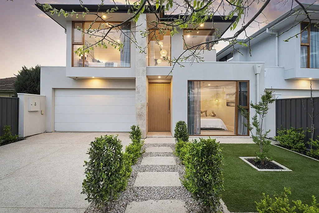 Modern two-story house with large glass windows, a wooden front door, a attached garage, and a landscaped front yard with bushes, small trees, and a concrete pathway.