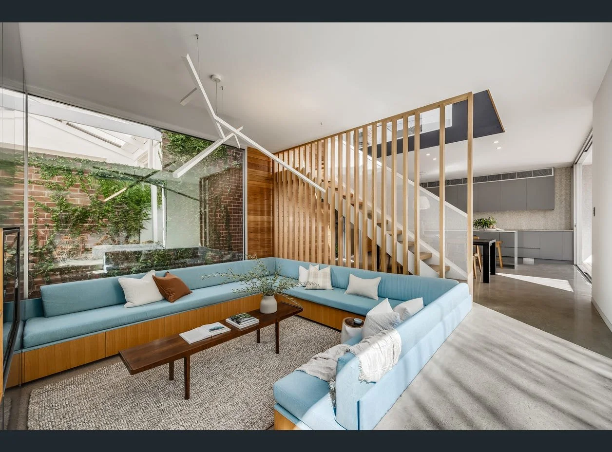 Modern living room with light blue sectional sofa, wooden coffee table, and large glass window showing outdoor brick wall and greenery.