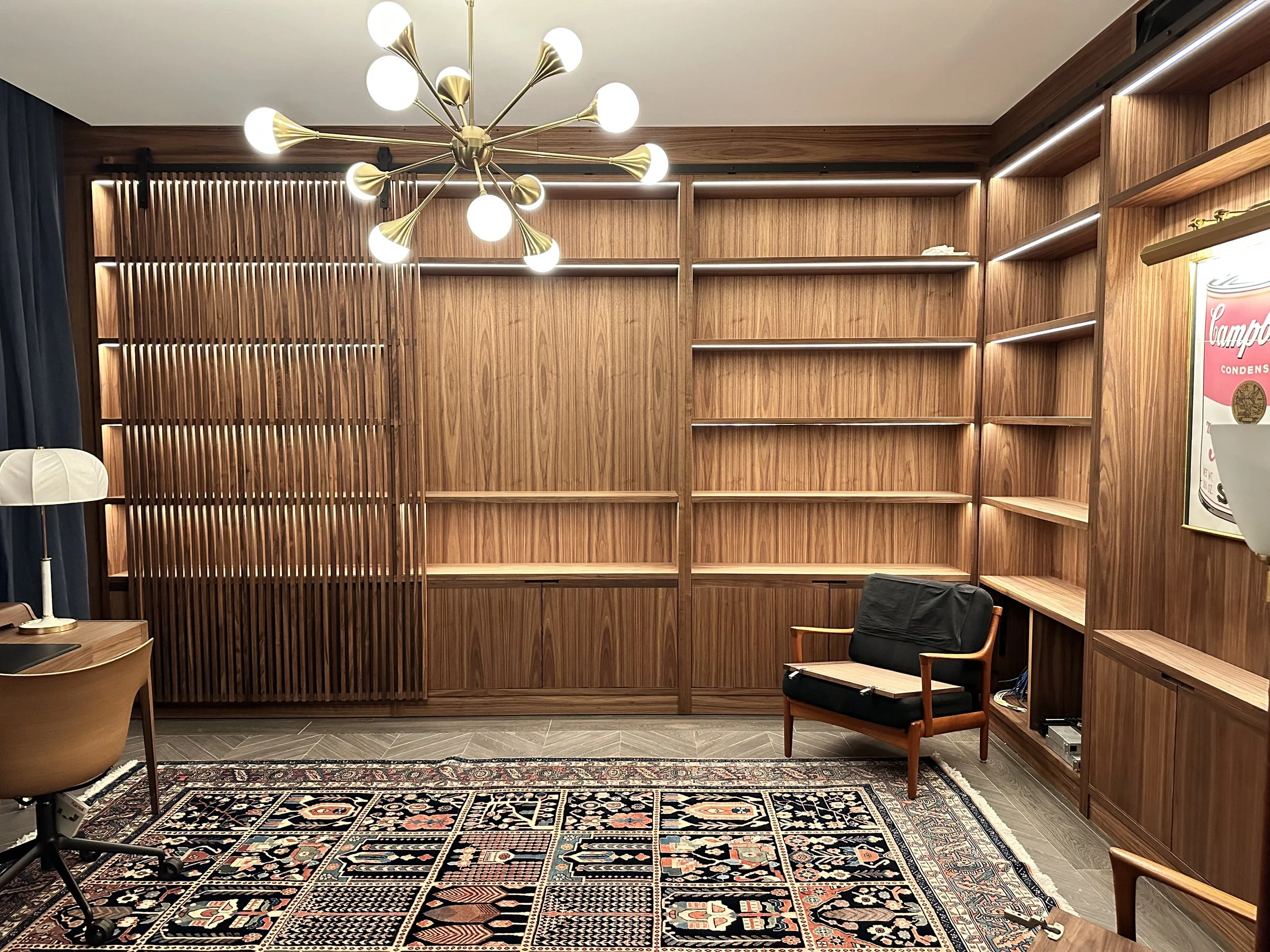 A room with wooden bookshelves, a vintage-style patterned rug, a modern chandelier with globe lights, a wooden chair with black cushions, and a desk lamp. The bookshelves are mostly empty, and part of a poster is visible on the right.
