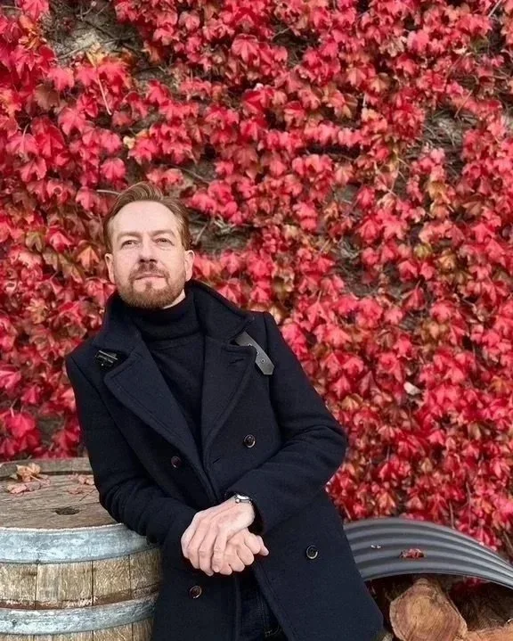 A man leaning against a wooden barrel with red and green ivy leaves in the background.