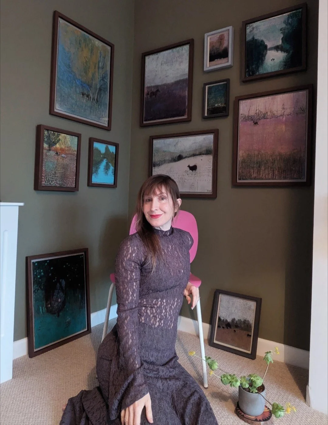 A woman with brown hair, wearing a long, sheer, dark lace dress, kneeling on the floor in a room with a gallery wall of framed landscape paintings and photographs. She is smiling at the camera, and there is a pink chair behind her and a potted plant nearby.