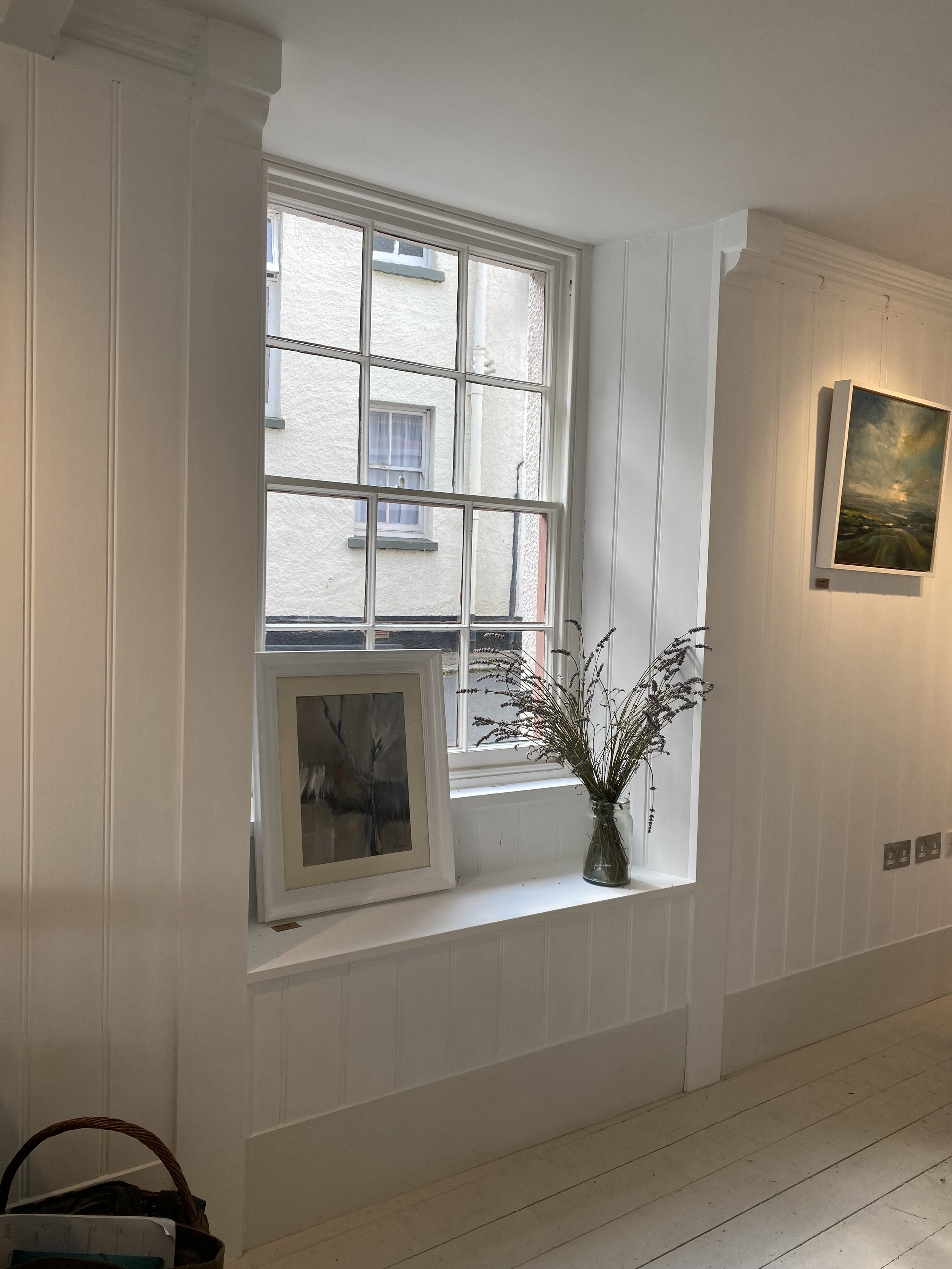 A white window ledge with a glass vase containing dried purple flowers, a framed artwork leaning against the wall, and a large window with multiple panes showing an outside building with white stucco walls and small windows.