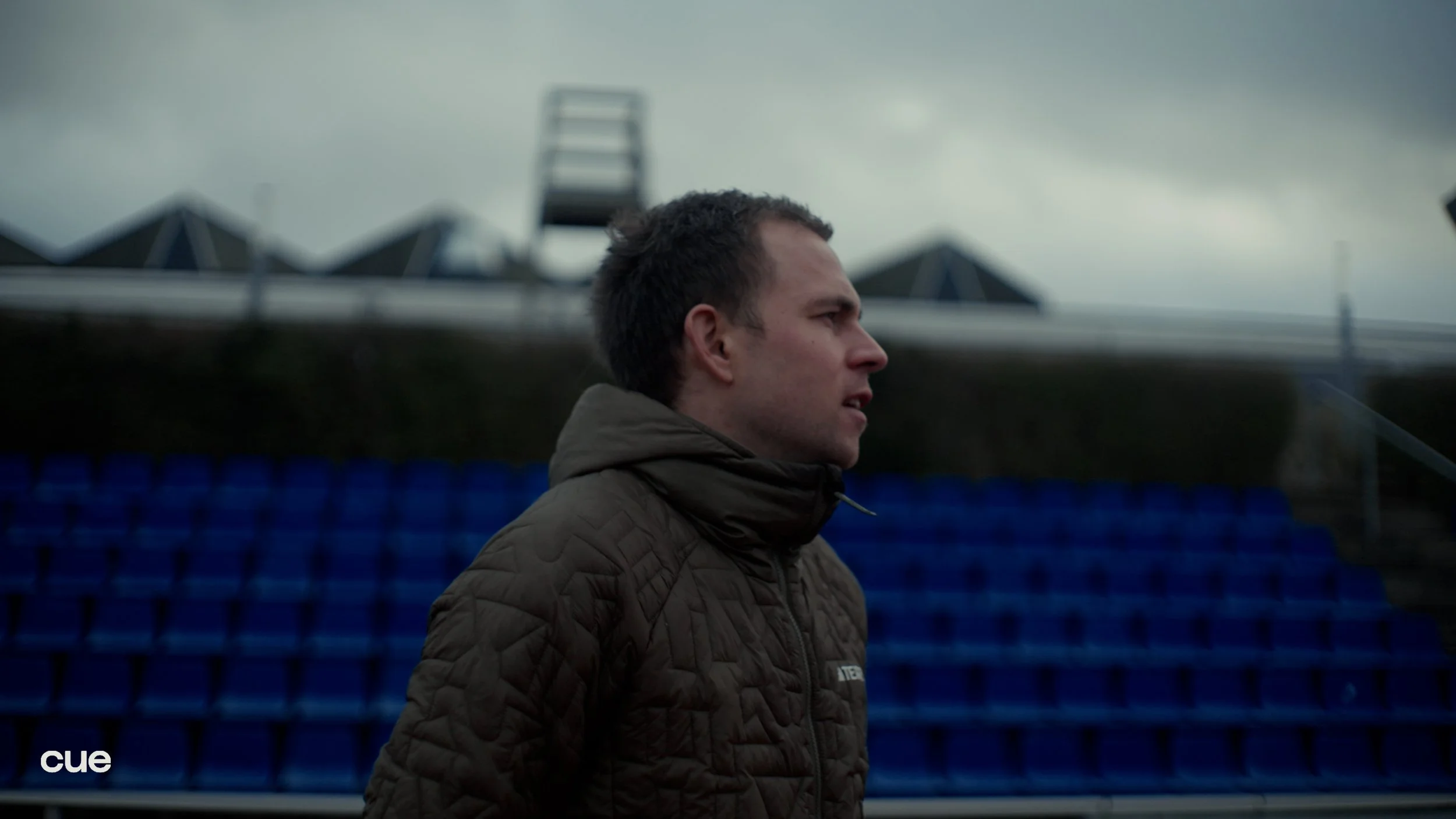 A man in a dark quilted jacket stands outdoors on a cloudy day, looking to the right. Blue stadium seats are visible behind him, with some roof structures and a ladder in the background.
