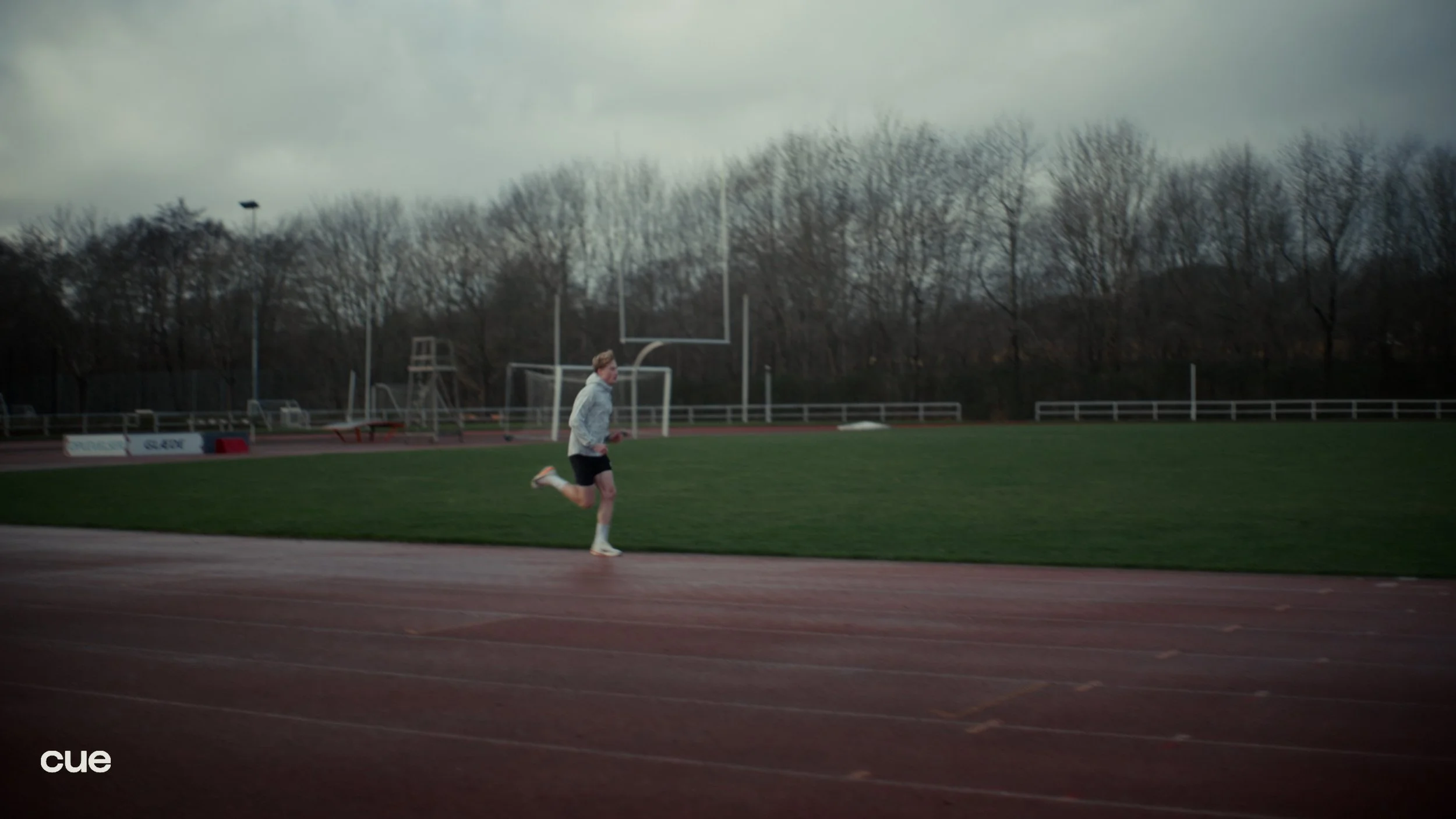 A person jogging on a track at a sports field with a running track, grass, and sports equipment in the background, under a cloudy sky.