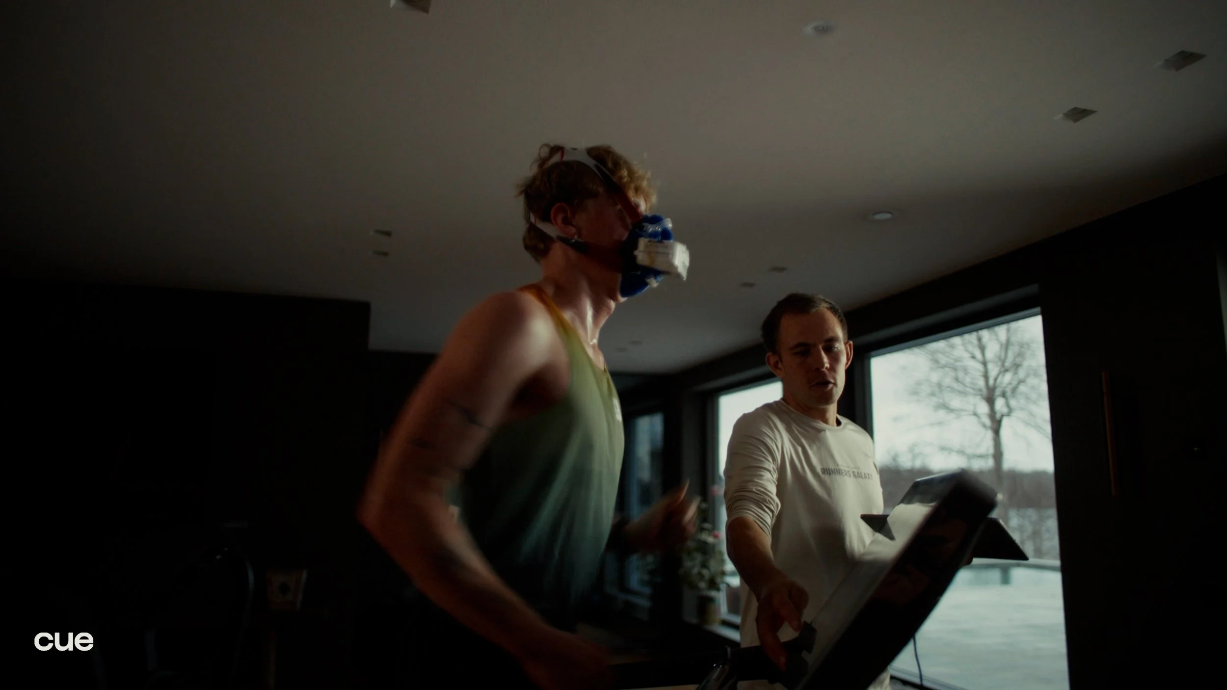 Two people indoors, one wearing a sleep apnea mask and running on a treadmill, and the other standing nearby holding a tablet, with large windows showing an outdoor scene with trees.