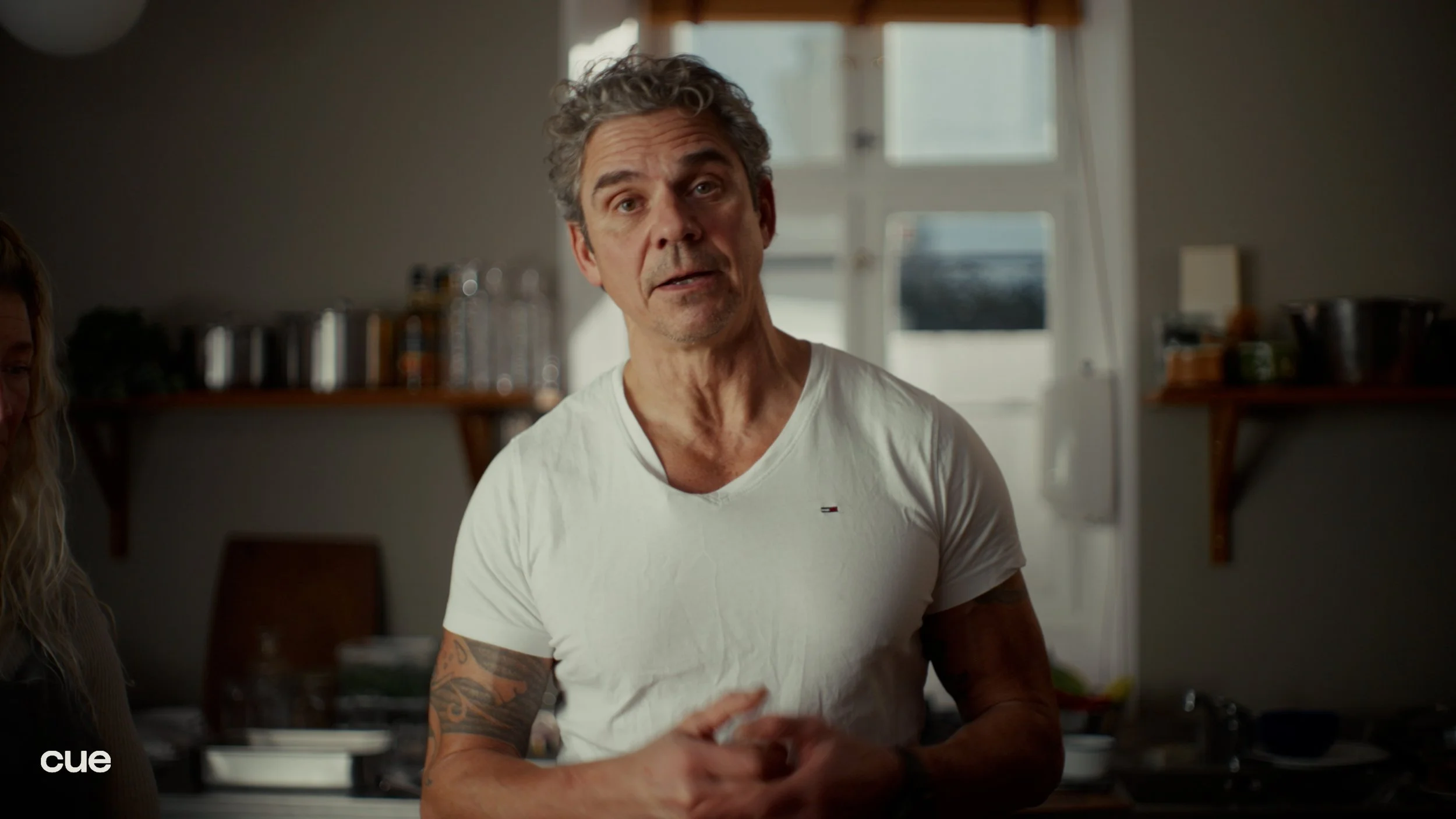 A middle-aged man with gray curly hair wearing a white t-shirt standing in a kitchen with shelves and a window in the background.