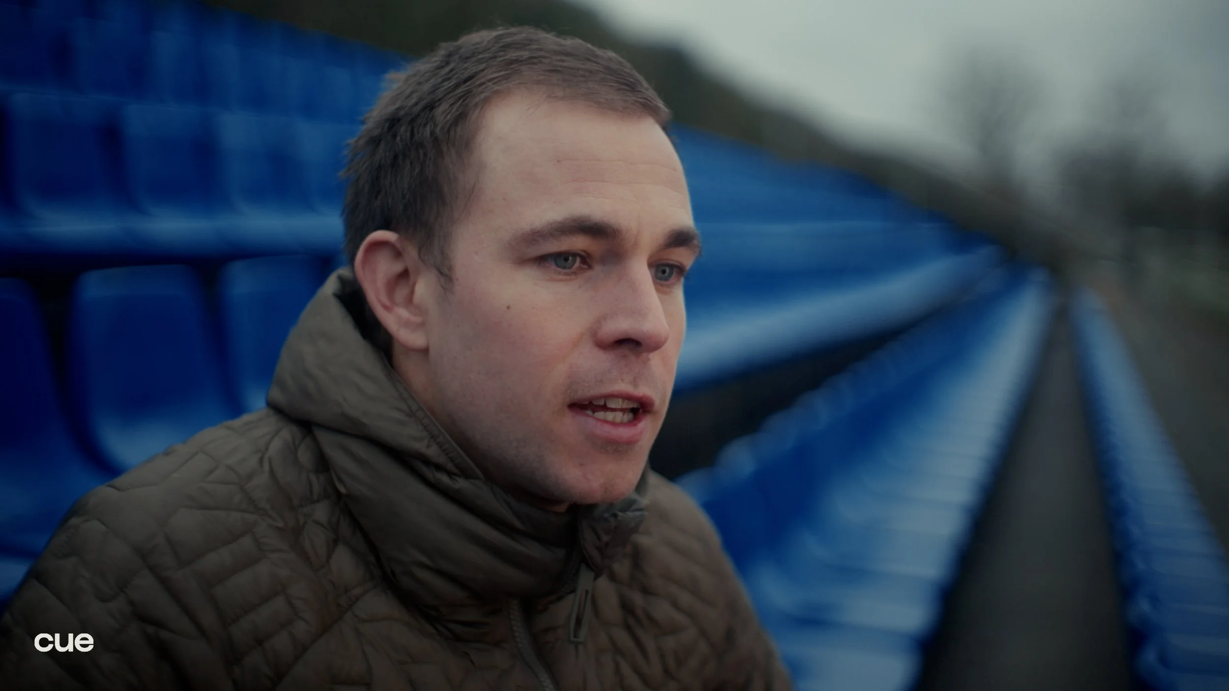 A young man with short brown hair and blue eyes, wearing a brown quilted jacket, is talking outdoors with blue stadium seats visible in the background.