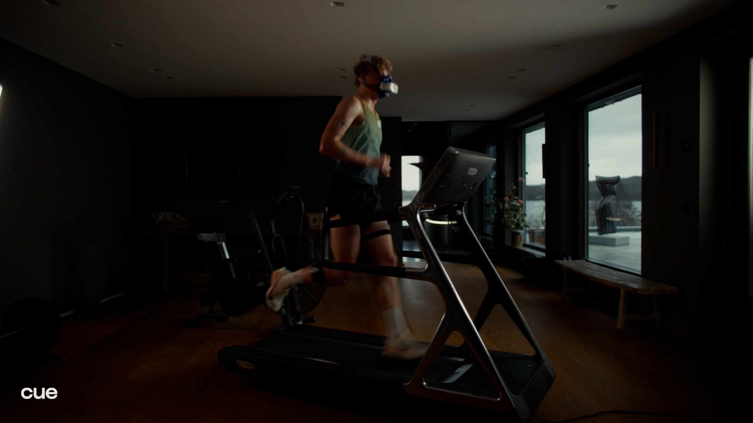 Person running on a treadmill in a dimly lit room with large windows, outside view of a balcony and outdoor furniture.