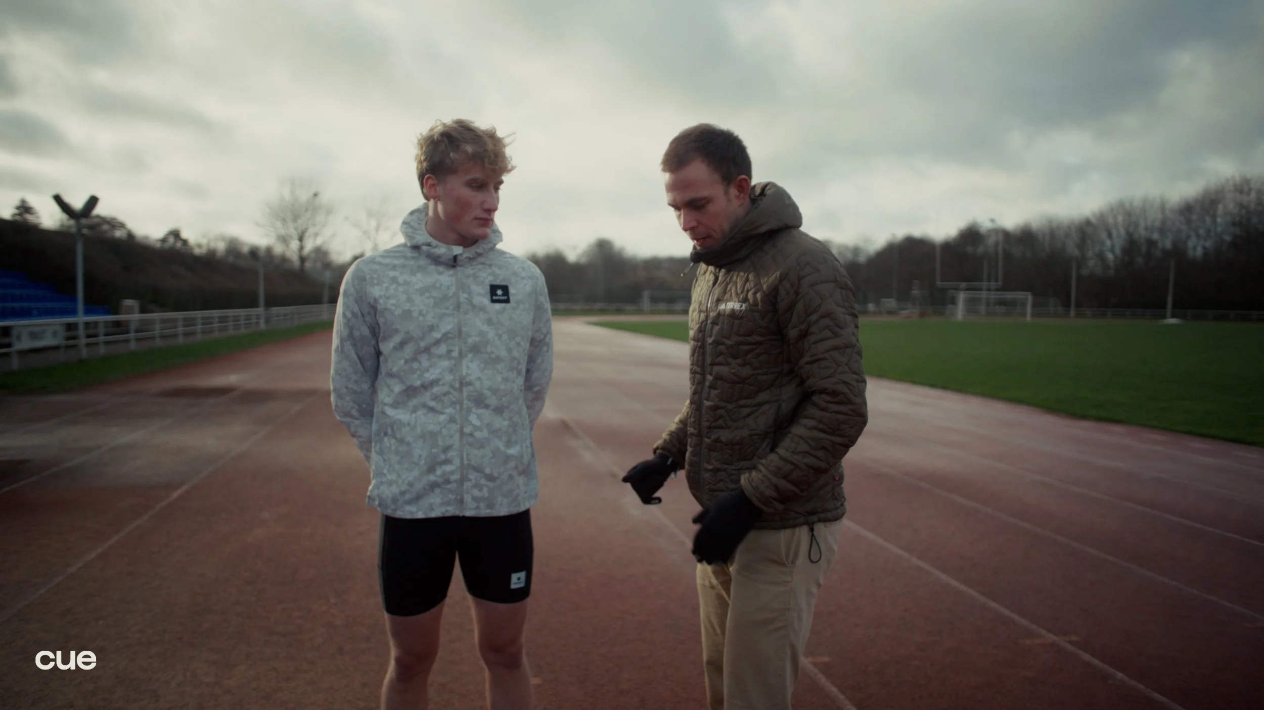 Two men standing on a running track, having a conversation. One is in athletic attire with a patterned jacket and shorts, and the other is in a brown quilted jacket and khakis. The background shows a cloudy sky and an outdoor sports field.