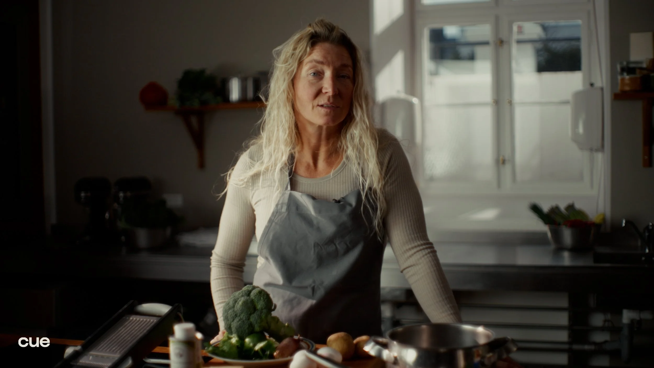 A woman wearing a gray apron standing in a kitchen with natural light from a window behind her, surrounded by vegetables and cooking tools on the countertop.