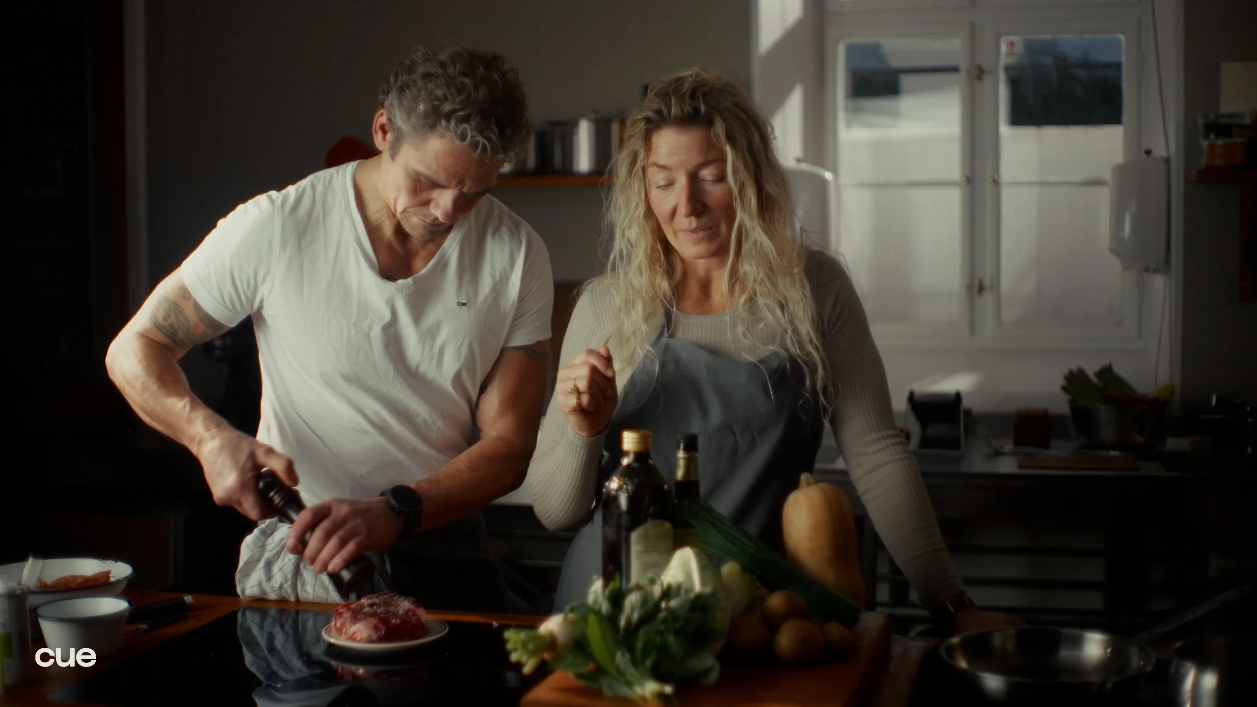 A man and woman preparing food together in a kitchen, with the man seasoning a piece of meat and the woman watching. The kitchen counter has various ingredients and bottles on it, and a window in the background lets in natural light.