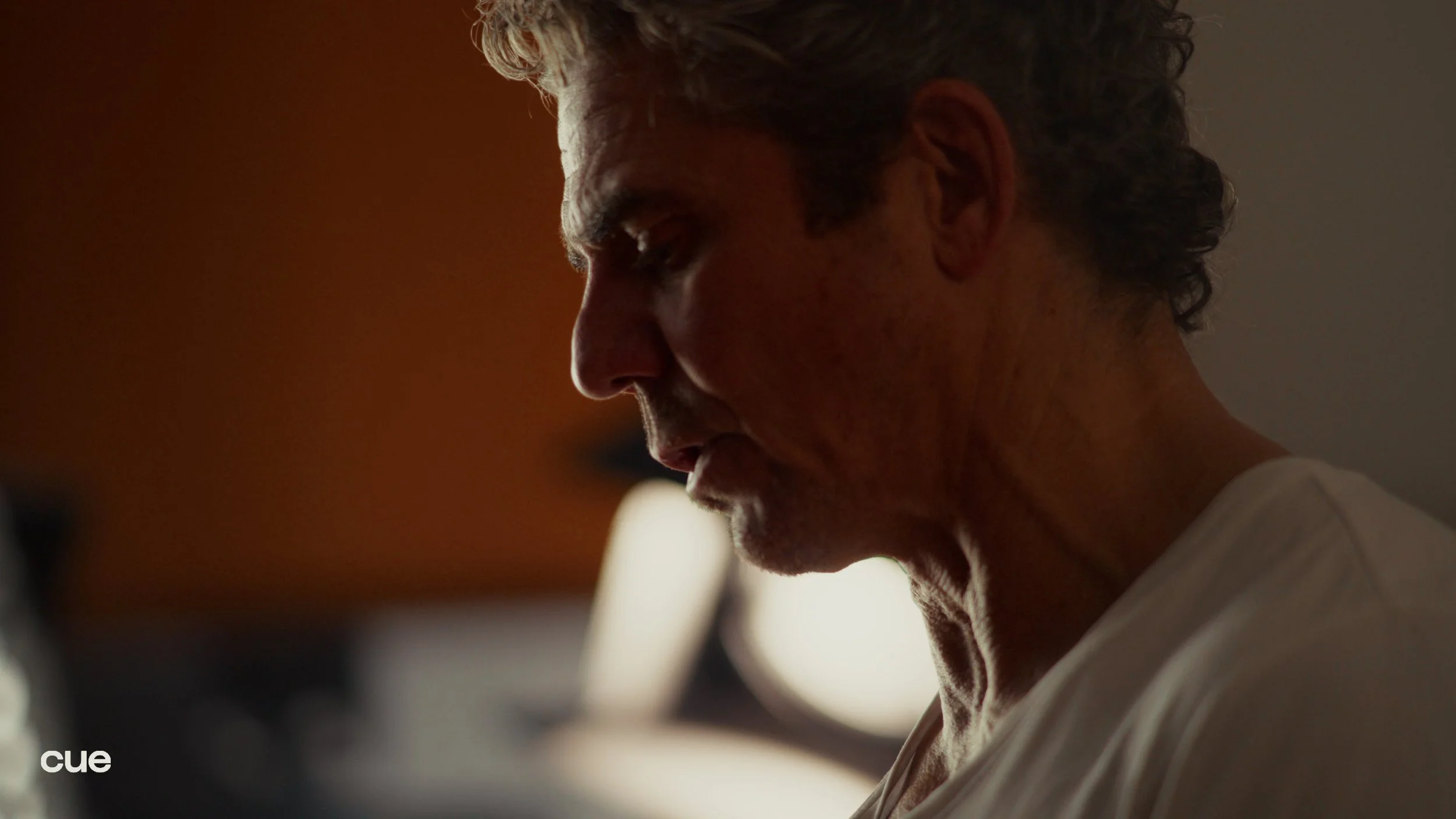 A side profile of an older man with gray hair, his head slightly bowed, in a dimly lit room.