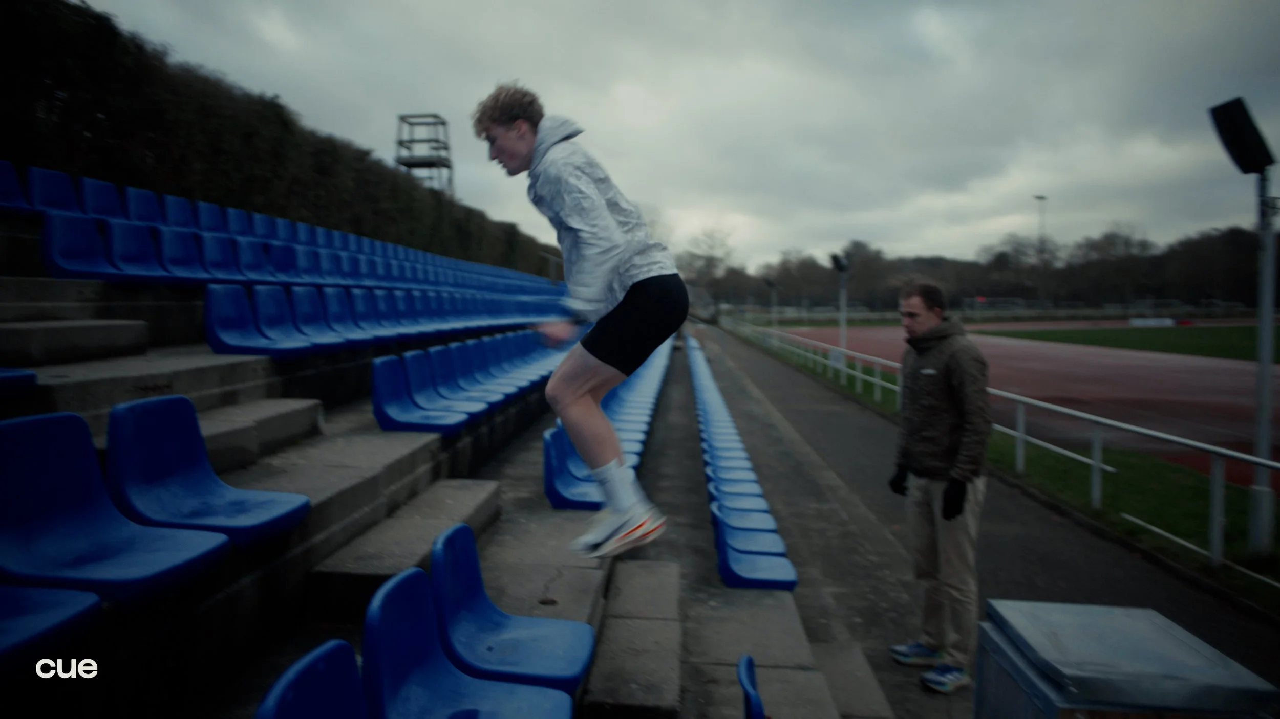 A person in athletic clothing jumping onto blue stadium seats on an outdoor track field as another person observes.