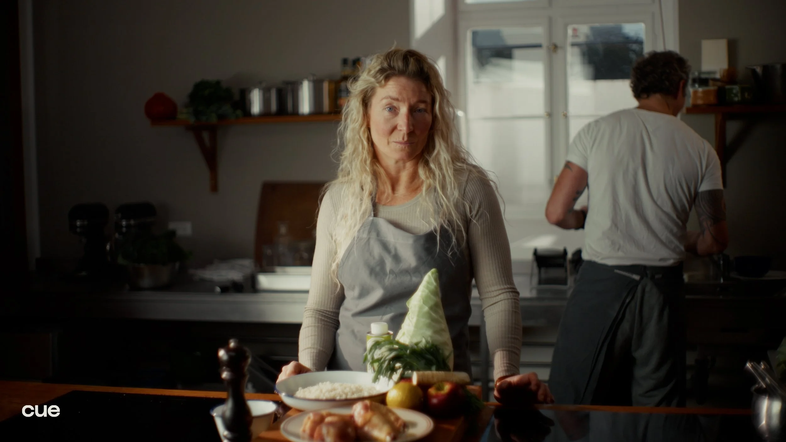 A woman with blonde curly hair wearing a gray apron with a green and white cheese sculpture in a kitchen. In the background, a man with tattoos on his arms is cooking. The kitchen has a modern design with a white window and shelves with jars and deco