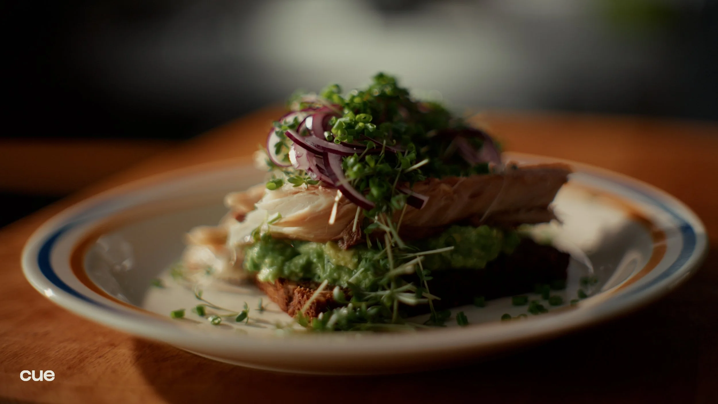 A plated dish of smoked fish topped with microgreens, onions, and a green sauce on a white plate with stripes.