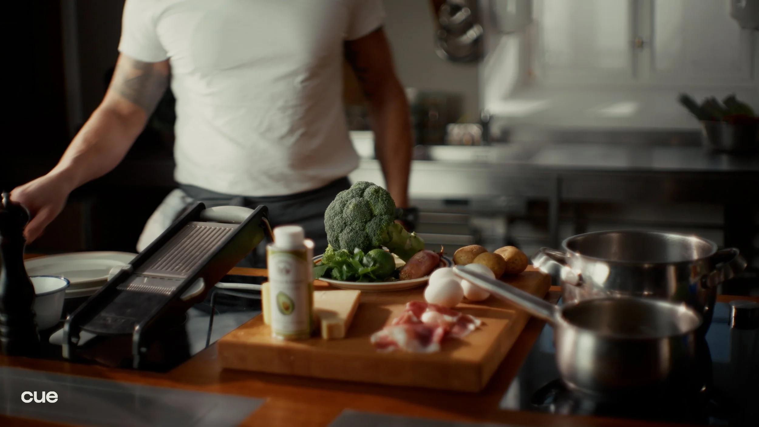 Kitchen counter with broccoli, potatoes, eggs, cheese, and bacon; stovetop and a person in a white shirt preparing food.