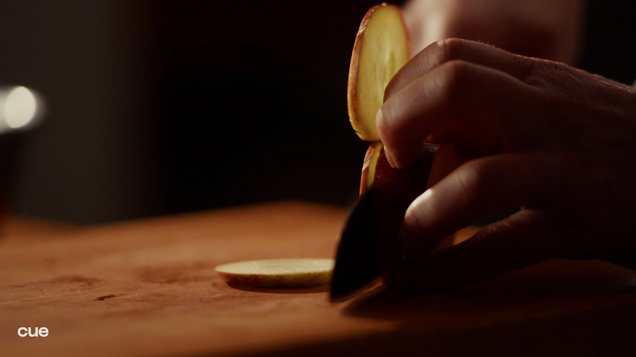 A person slicing a red apple on a wooden cutting board in a dimly lit setting.