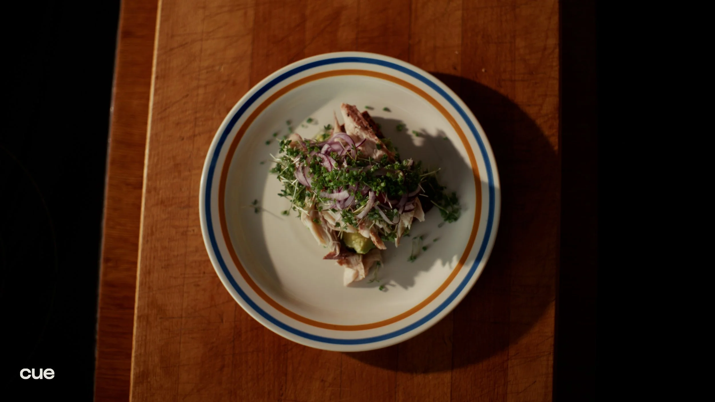 A plate with shredded cooked chicken, sliced red onions, microgreens, and sliced avocado, on a wooden table.