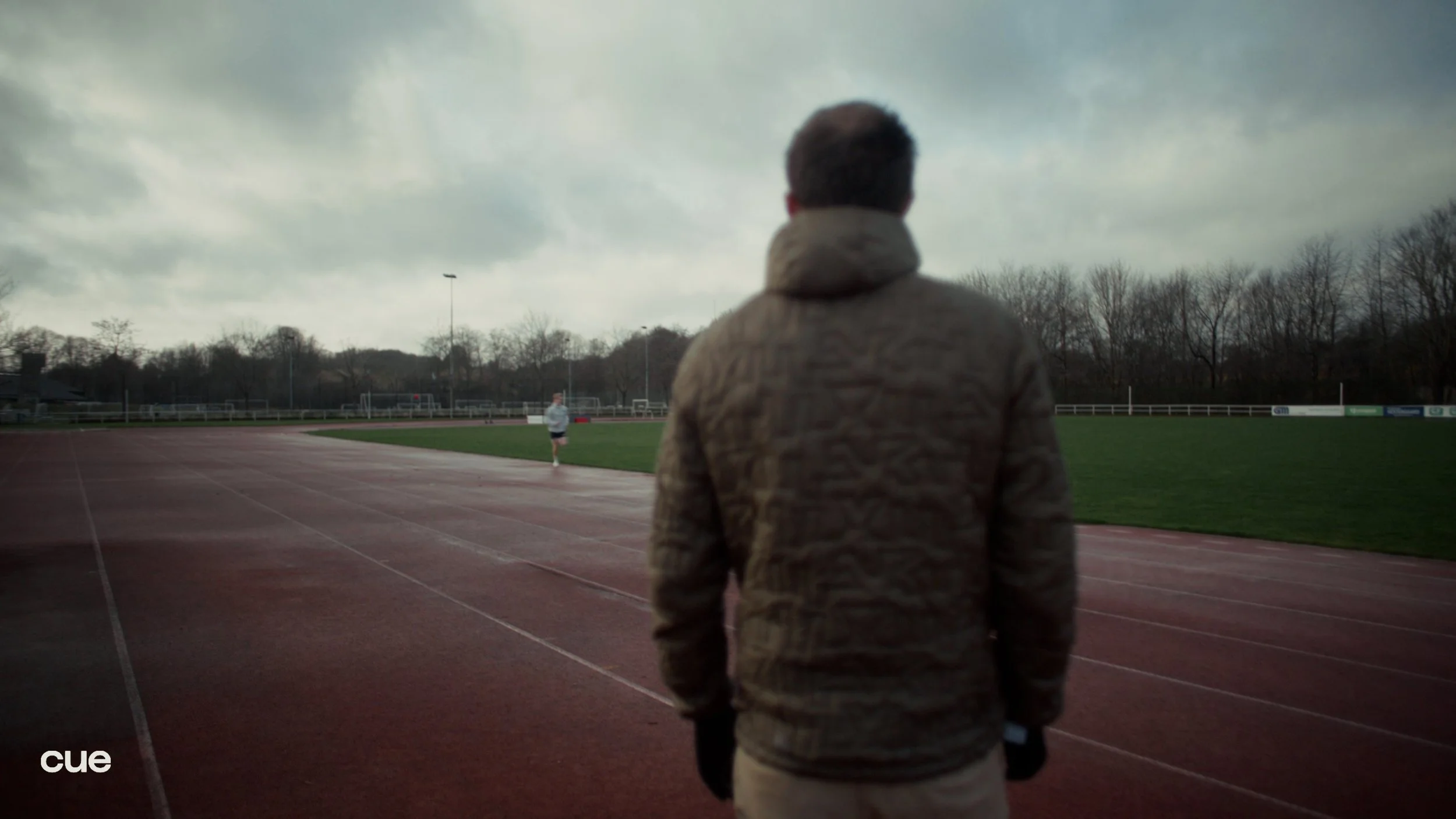 A person wearing a beige jacket stands on a running track, watching a person running on the track in a sports field under cloudy skies.